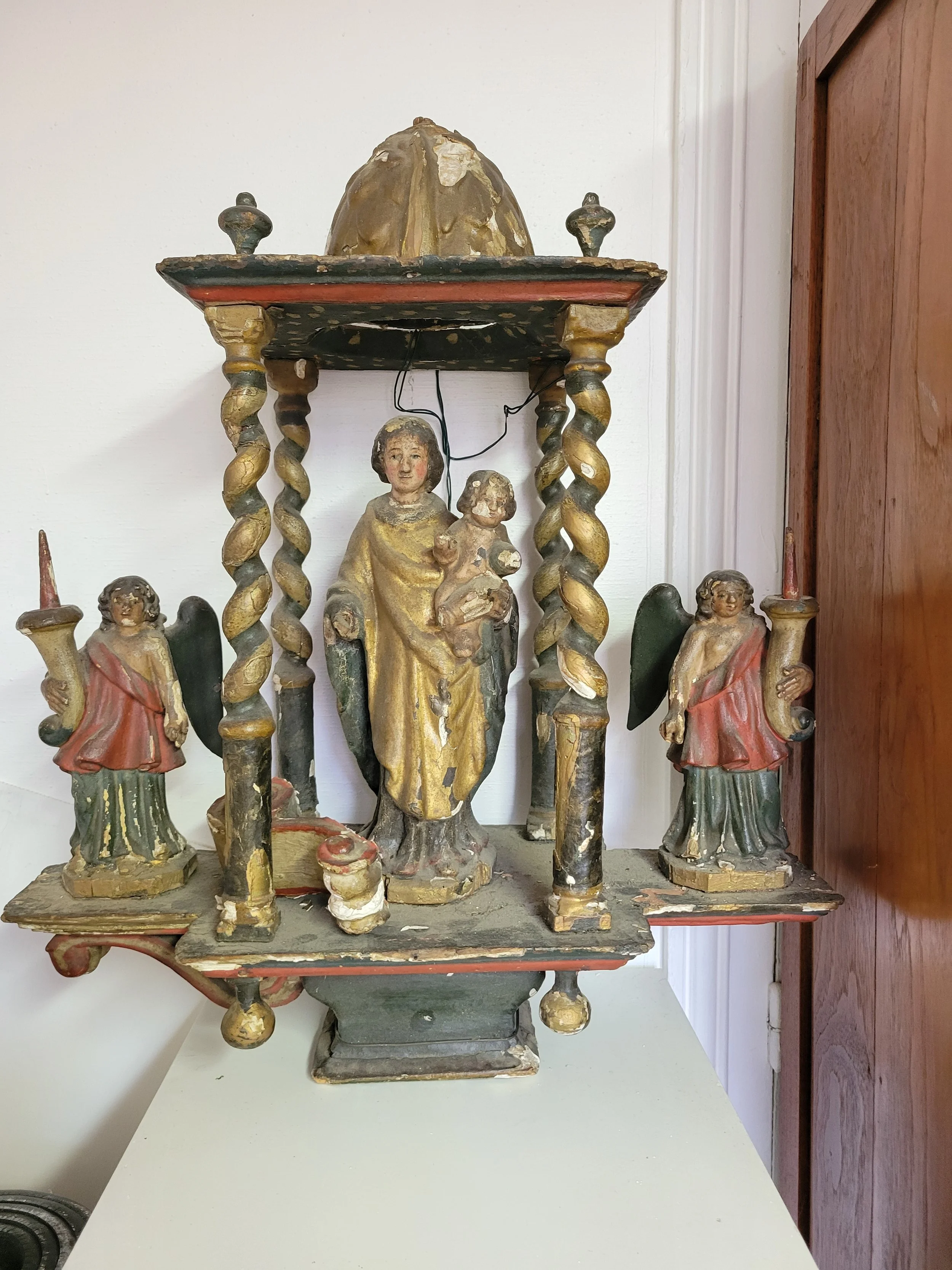 Une antique sculpture religieuse représentant la Vierge Marie avec l'enfant Jésus, entourée d'anges, en bois peint et doré, montrant des signes d'usure.