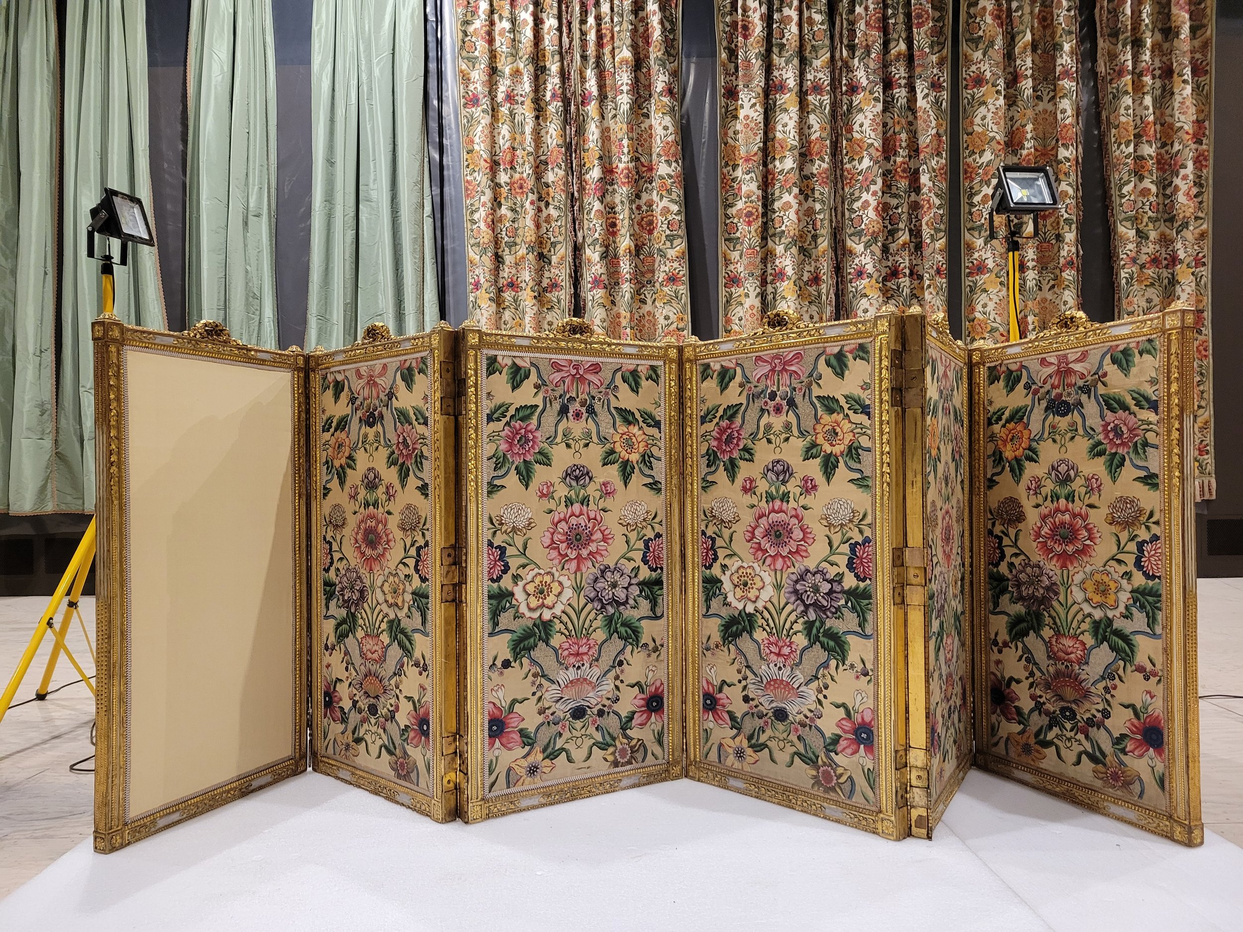 Ecran de confidentialité décoré de panneaux avec motifs floraux, dorés, sur fond beige, et drapeaux de fond avec des rideaux à motifs floraux.