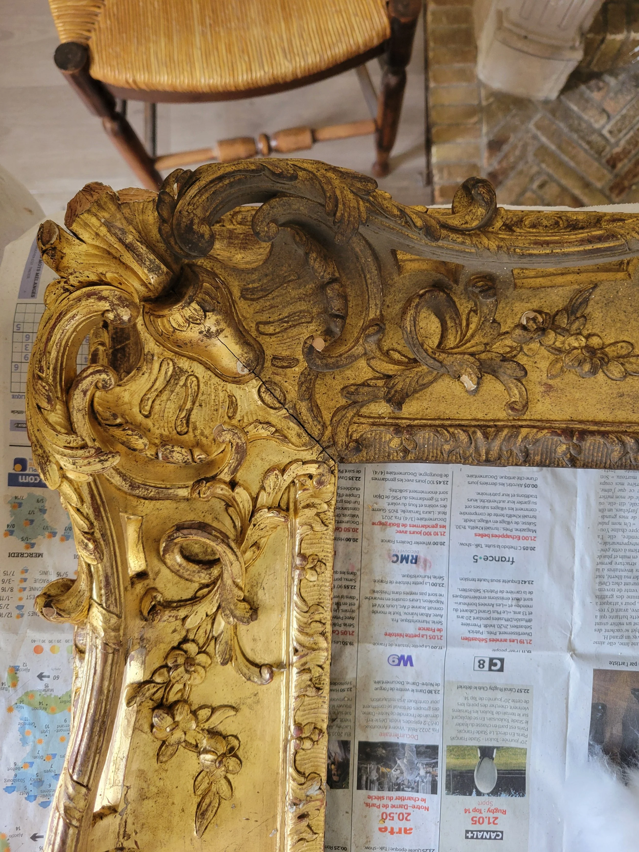Détail d'une sculpture décorative en bois doré avec des motifs floraux et feuillages, posée sur une surface recouverte de journaux.