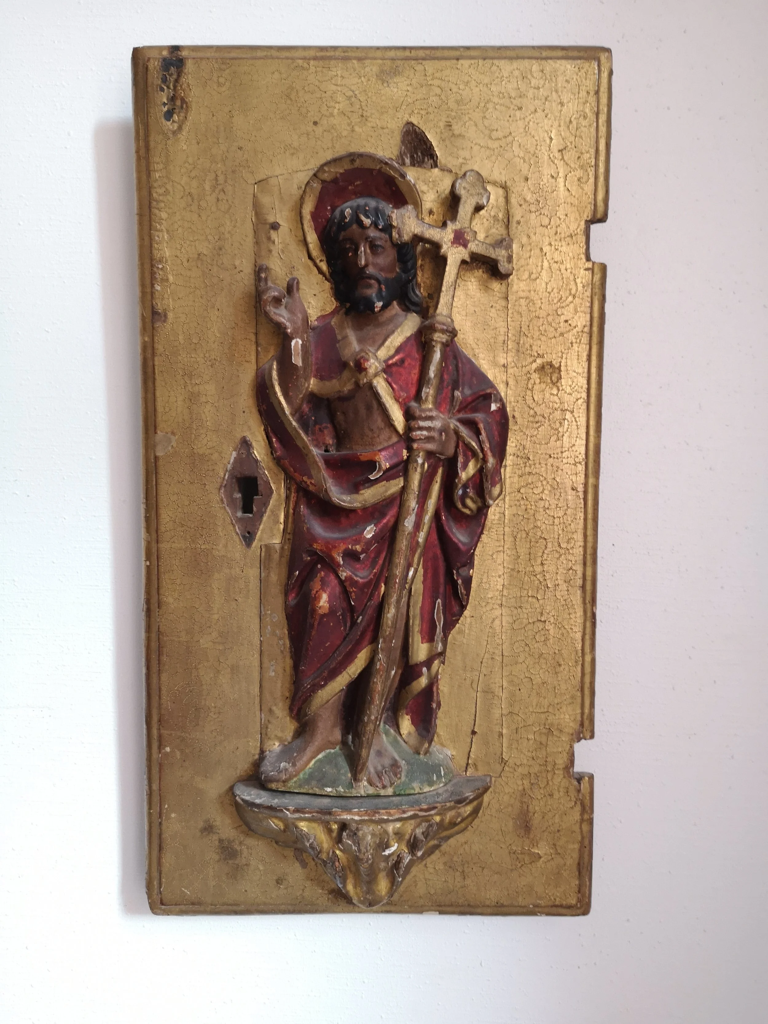 Une sculpture religieuse en bois représentant Jésus-Christ avec une couronne d'épines, tenant une croix, sur un fond doré. La sculpture est encadrée et semble ancienne, avec des détails peints en rouge, noir et or.
