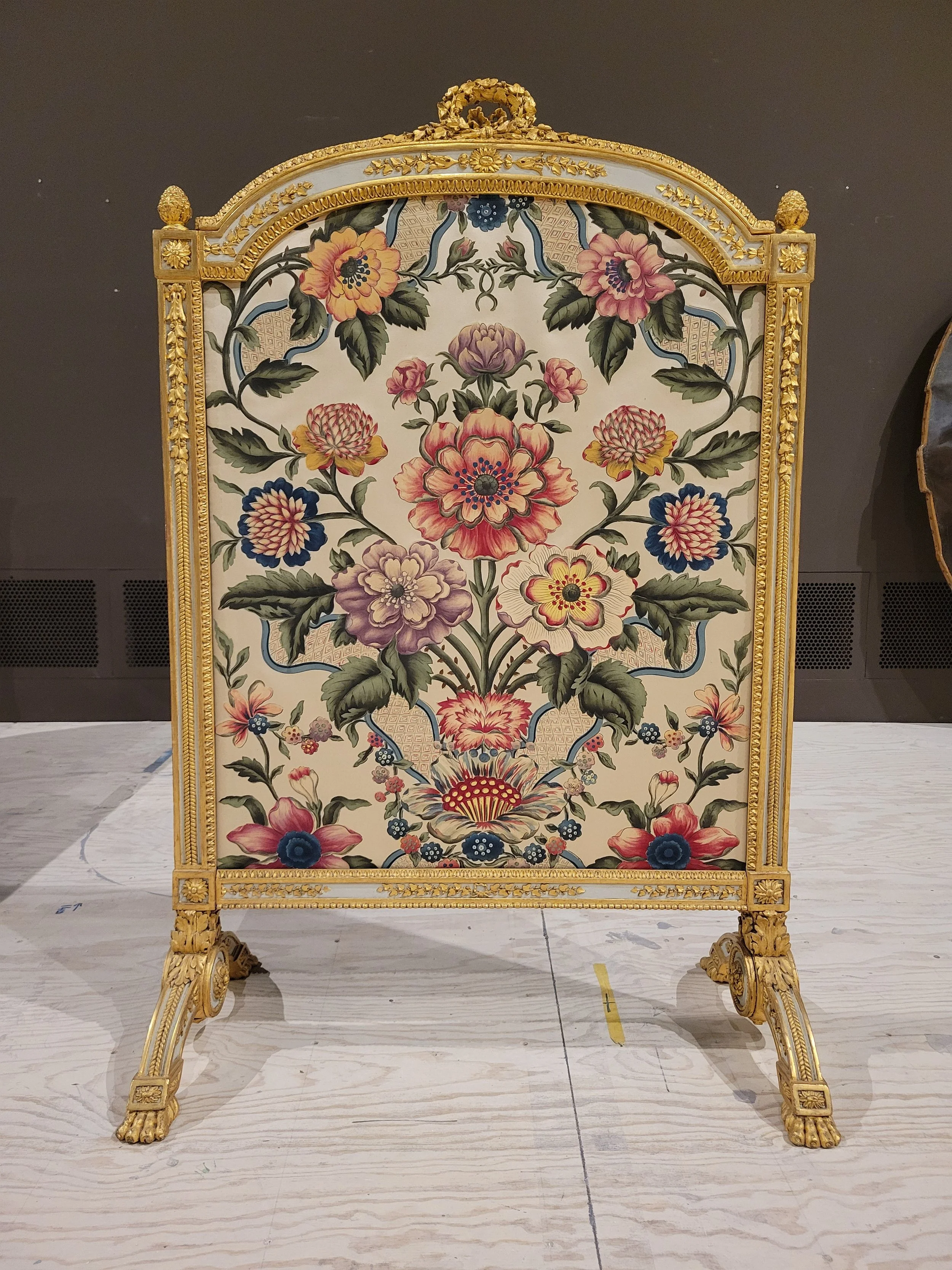 Un fauteuil ornée de fleurs peintes avec un cadre en or détaillé, posé sur un sol en bois clair dans une galerie ou un musée.