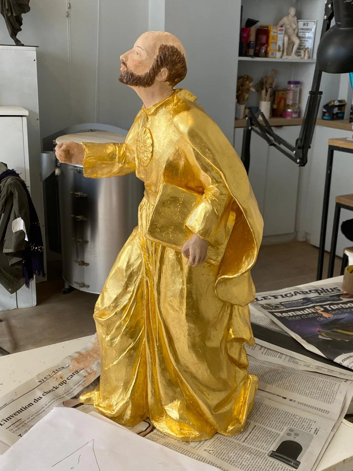 Statue représentant un homme avec une barbe, vêtu d'une tunique dorée, tenant un livre dans la main. Atelier avec étagères et objets divers en arrière-plan.