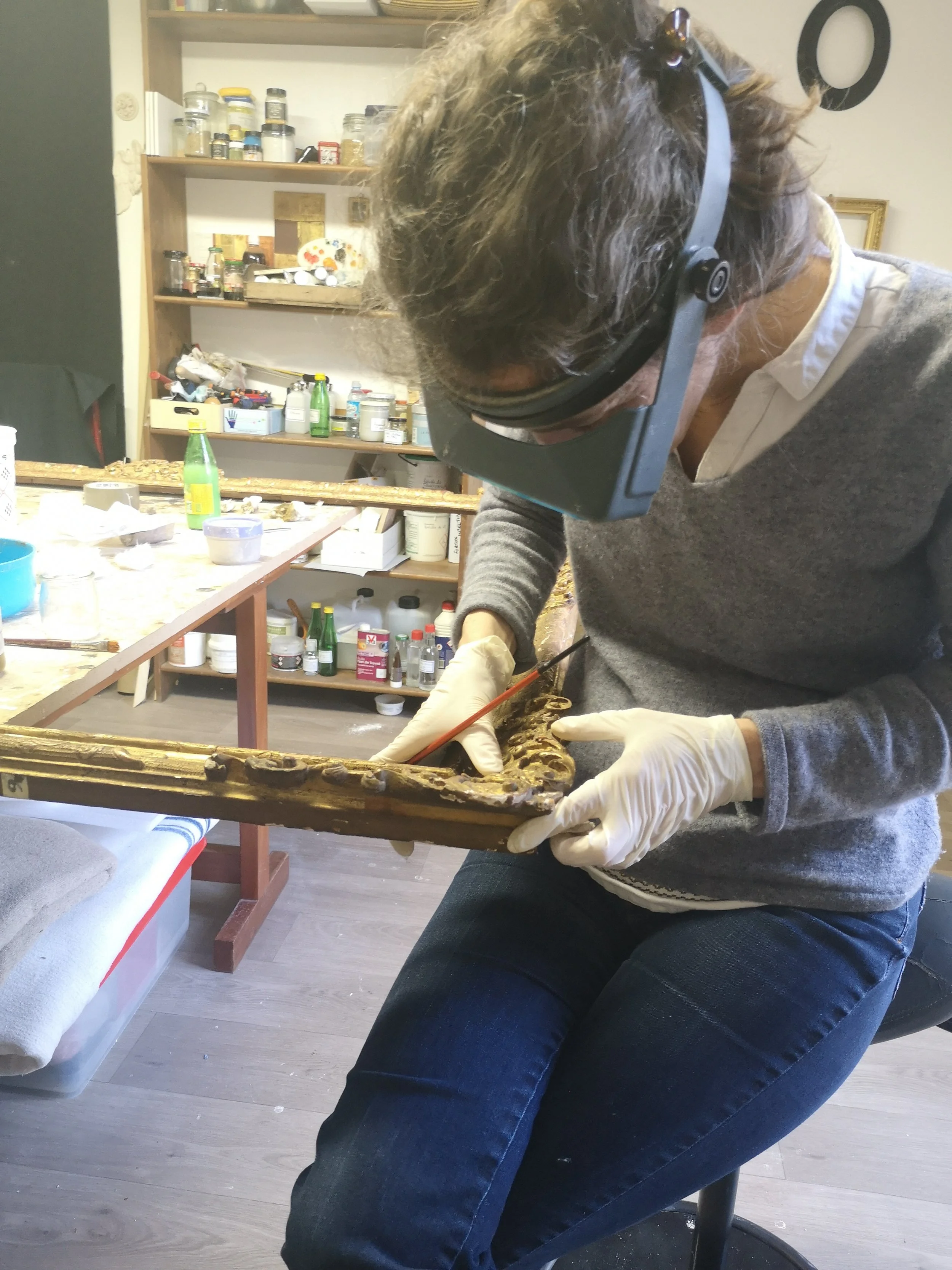 Une personne portant des gants, un casque de protection, un masque et un pull gris, nettoie ou restaure un cadre en bois doré. La pièce semble être un atelier avec des étagères remplies de bocaux, de tubes et de produits à l’arrière, et une table de travail devant.