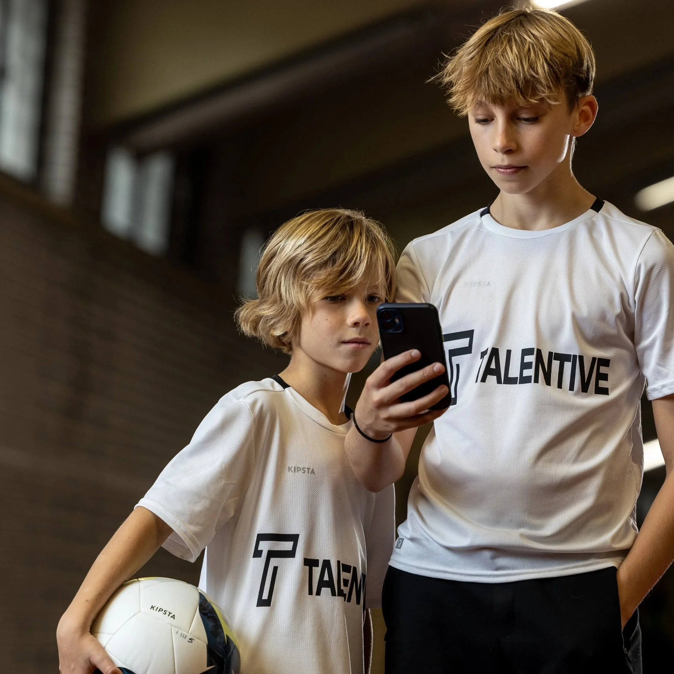Two boys in sports jerseys standing in an indoor sports facility looking at a cellphone, one holding a soccer ball.