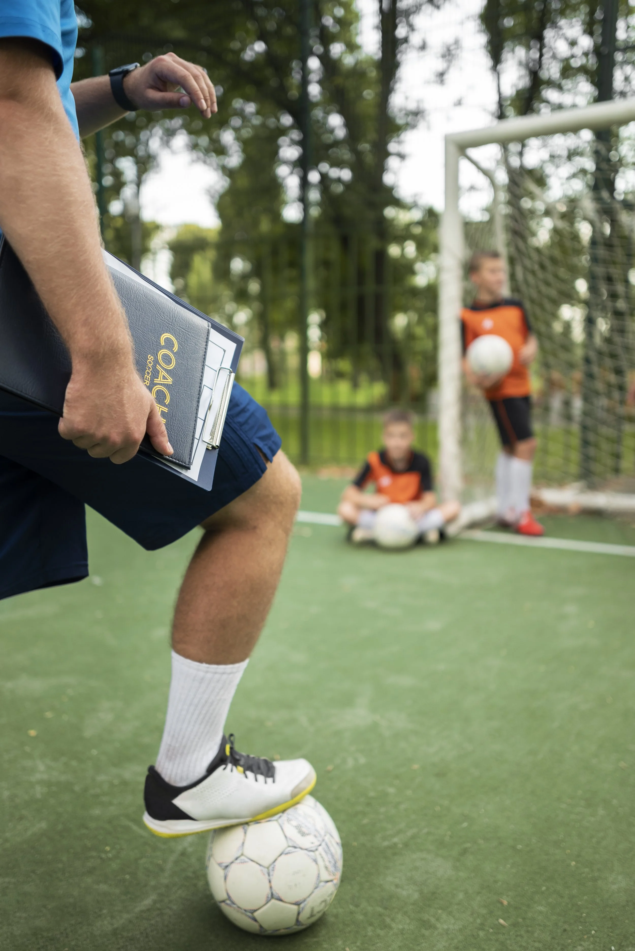 Soccer coach prepares to demonstrate a soccer move, with a soccer ball under one foot, while two young soccer players, one sitting and the other standing with a ball, wait near the goal on an outdoor field surrounded by trees.