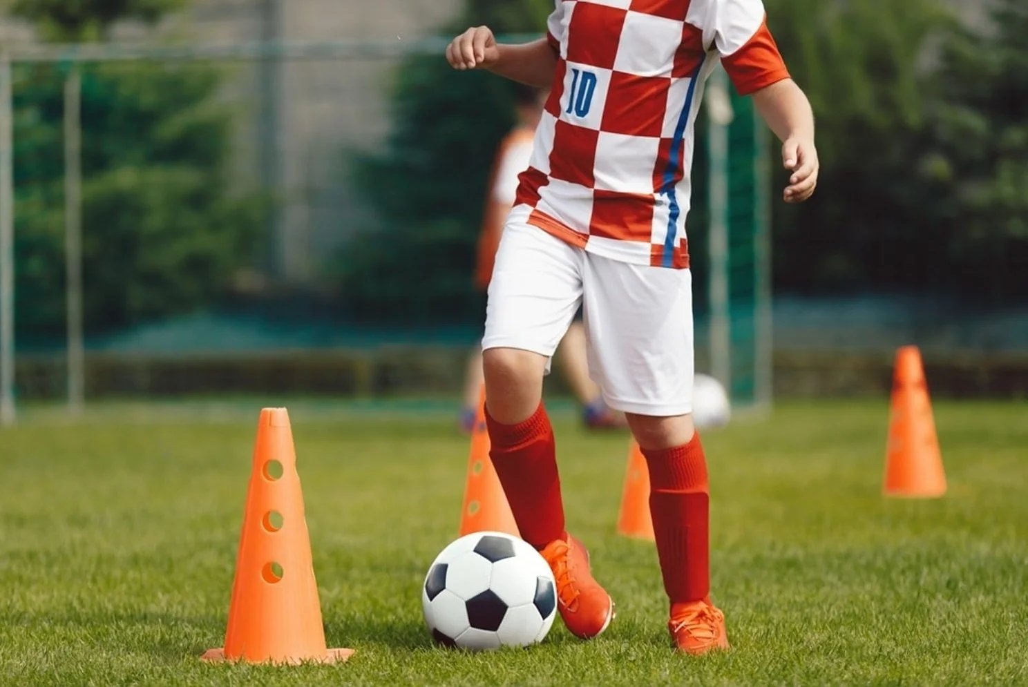 Child in red and white soccer uniform practicing dribbling around orange cones on a soccer field.