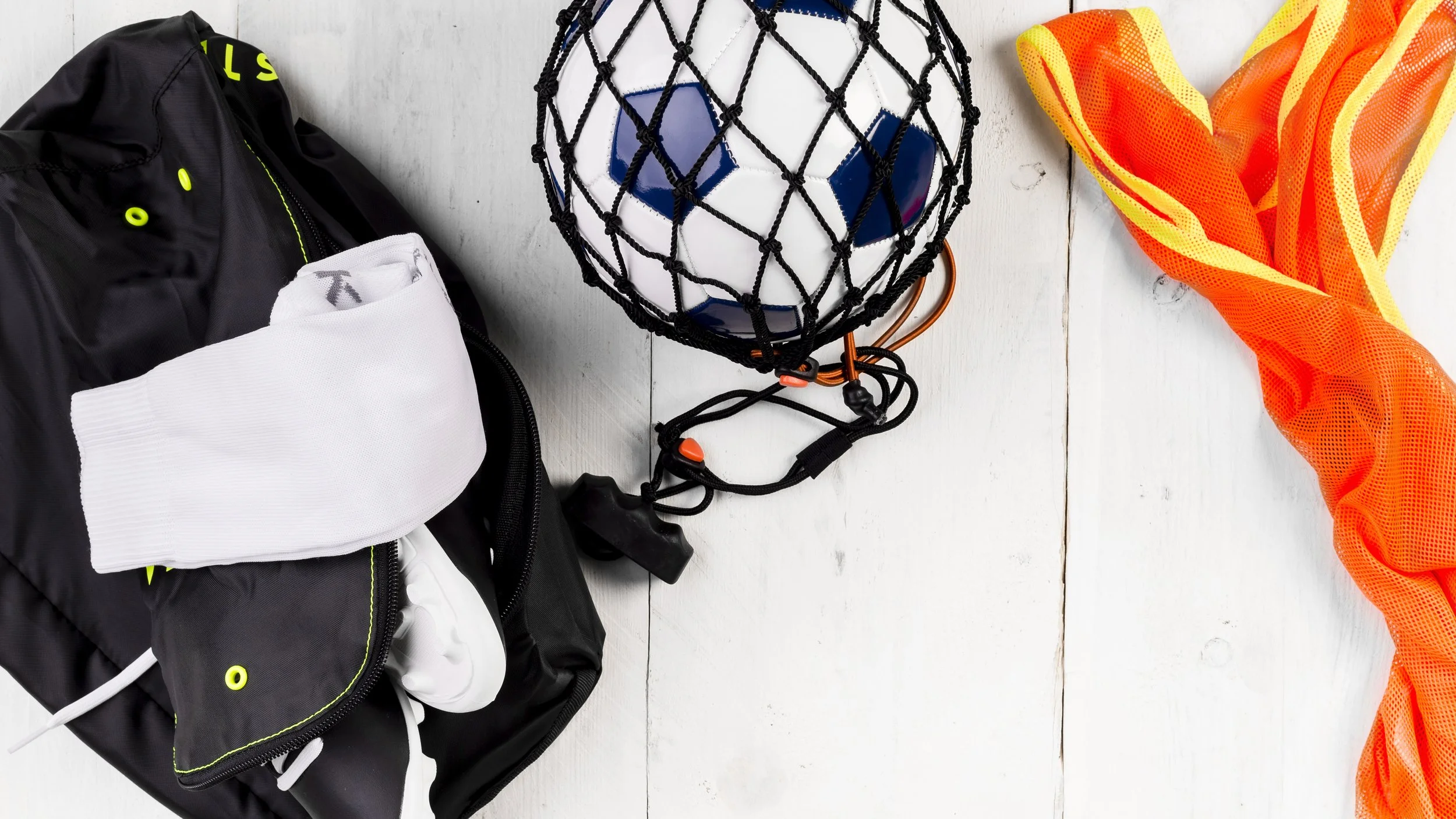 Soccer ball in a net, black backpack, white socks, and an orange and yellow mesh sports vest on a white wooden surface.
