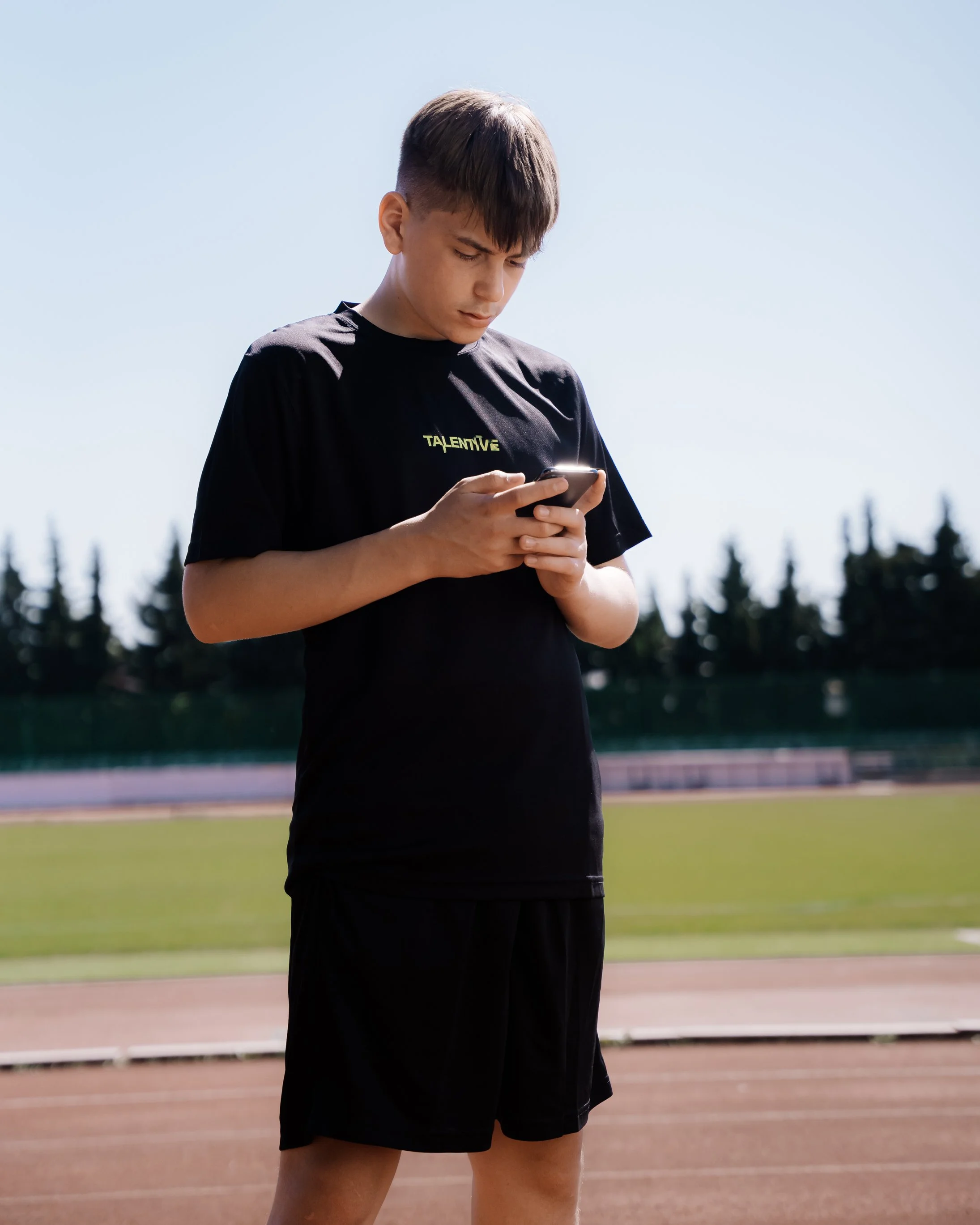 Young man in black sportswear standing on an outdoor track, looking at his phone, with a grassy field and trees in the background.
