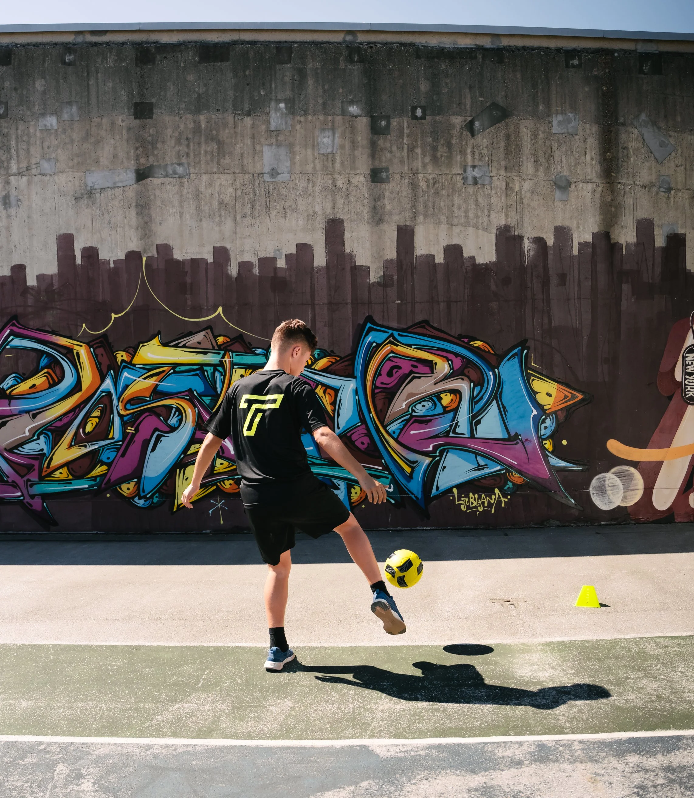 A boy playing soccer on a court with a graffiti wall in the background.