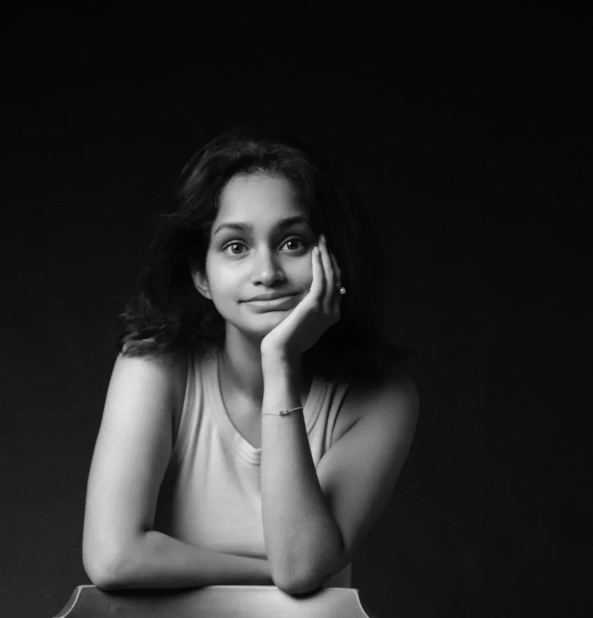 Black and white portrait of a young woman with short wavy hair, resting her face on her hand, smiling softly, against a dark background.