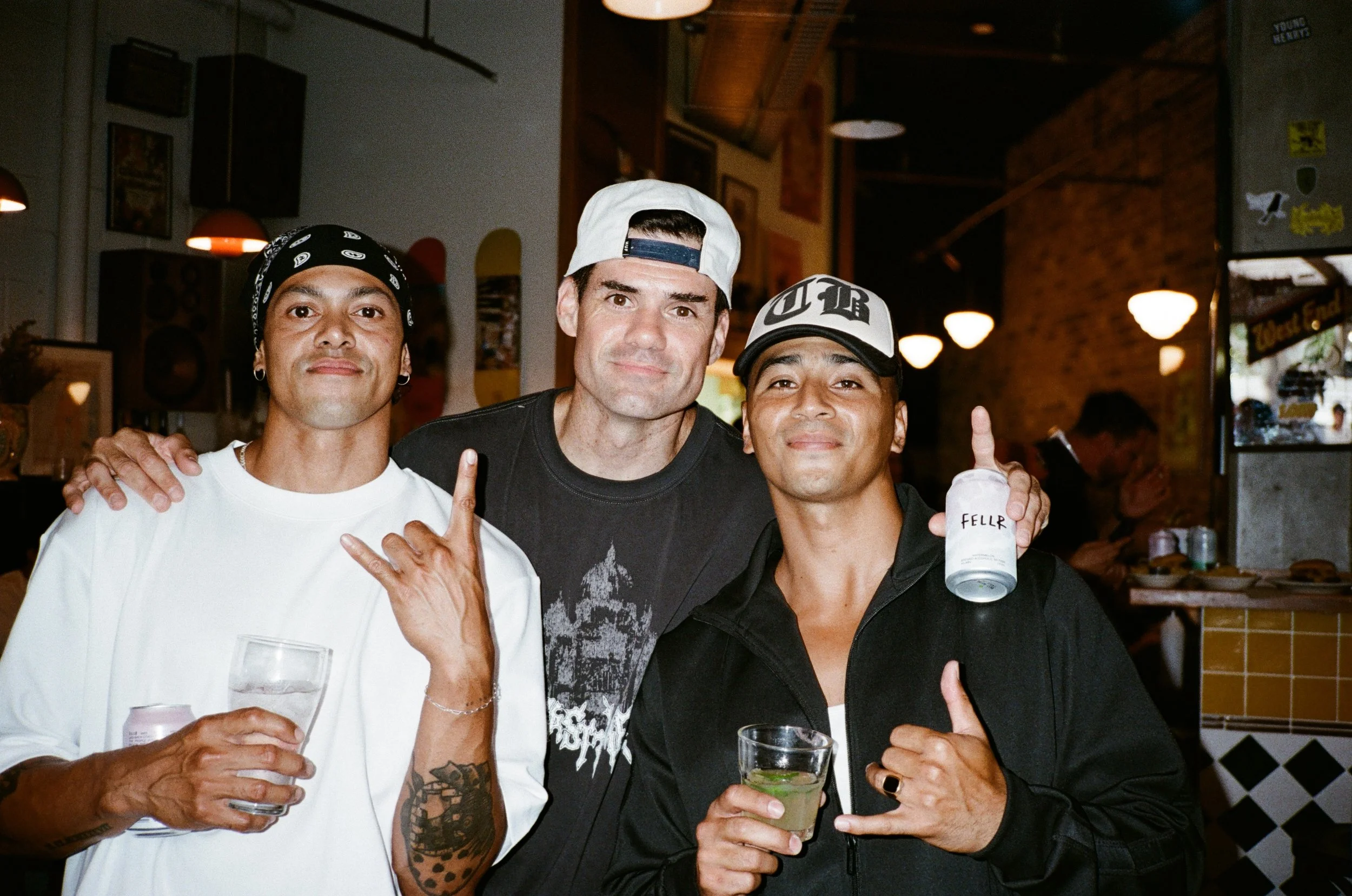 Three men posing together at a bar or restaurant, smiling and holding drinks, with a brick wall and dim lighting in the background.