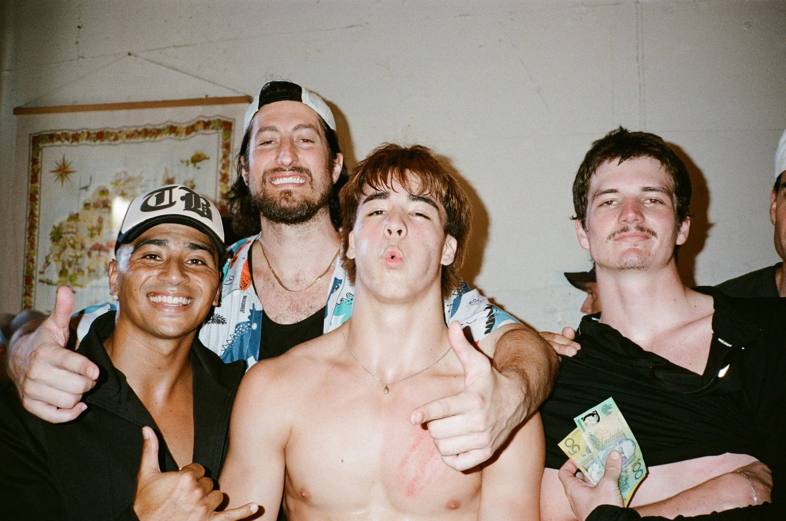 A group of five young men, shirtless and wearing casual clothing, celebrating together indoors. They are smiling, some giving thumbs up, and one person is holding Australian currency notes. The background features a decorated wall hanging.