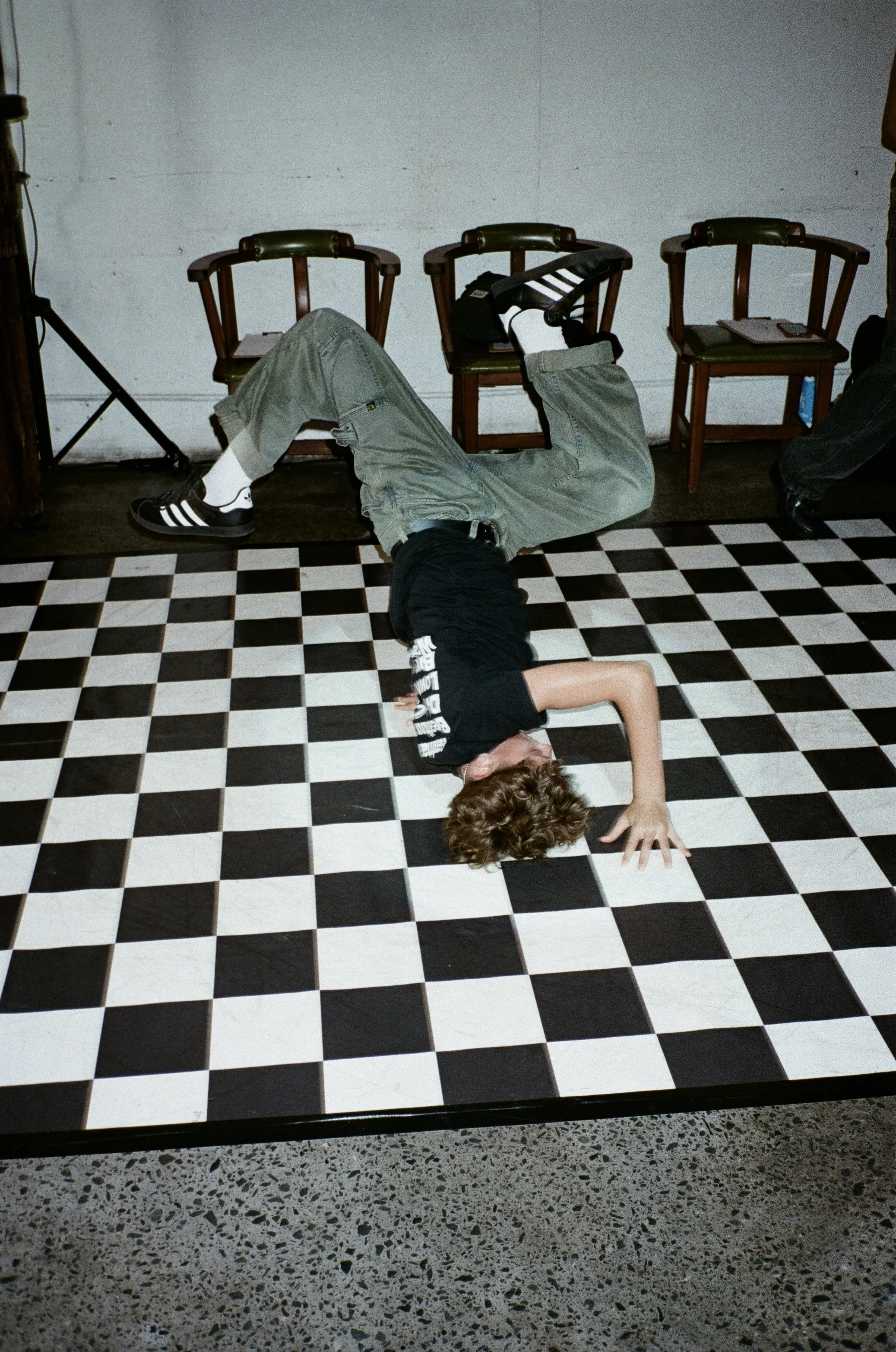 Person lying on checkered black and white tile floor, with one arm reaching up and the other resting on the floor, legs crossed, and head turned to the side. Four empty chairs are lined up against a white wall in the background.