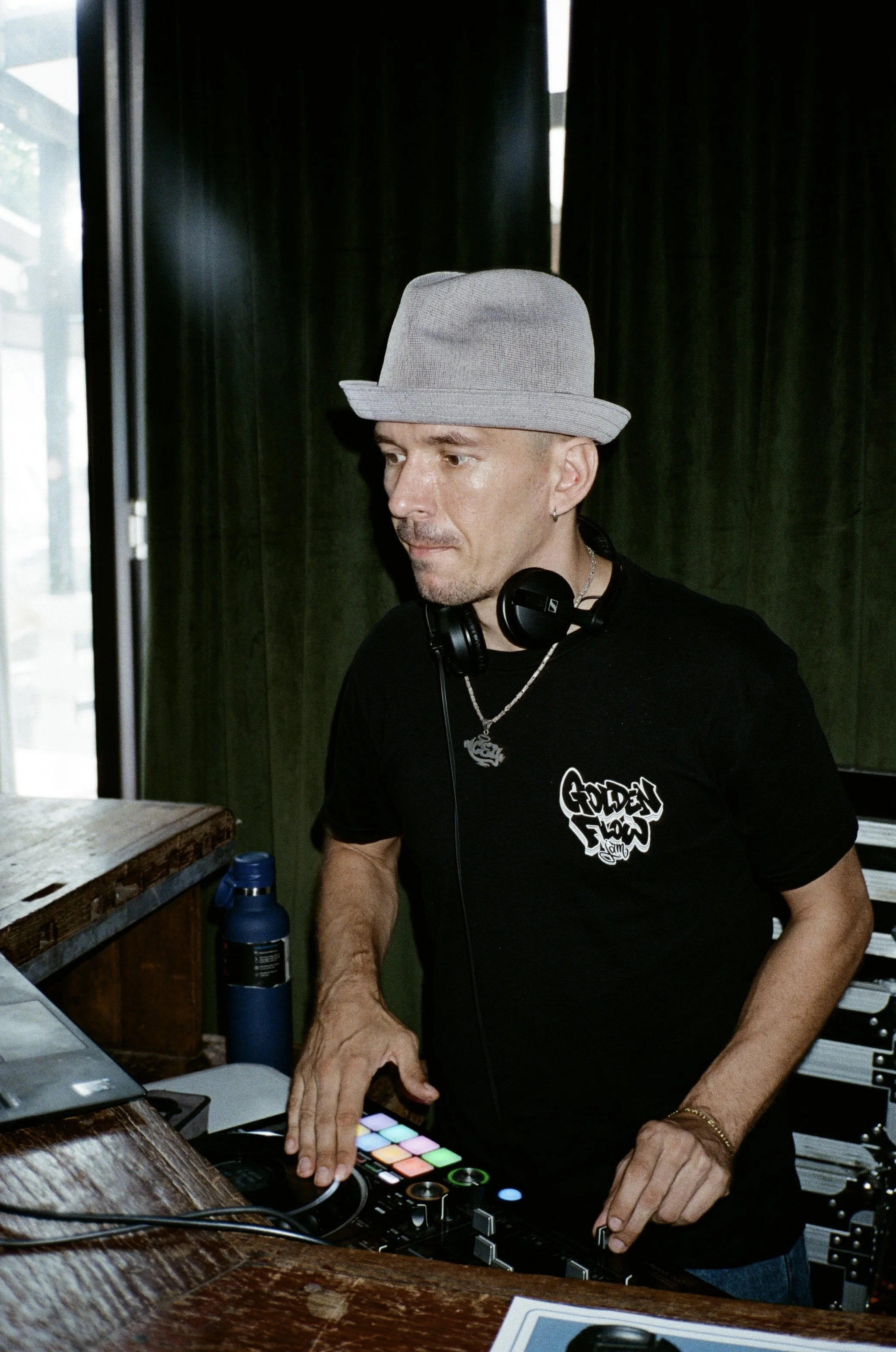 Man DJ wearing a light gray fedora hat and black t-shirt with a graphic text, using a DJ controller with colorful pads, in a dimly lit indoor setting.