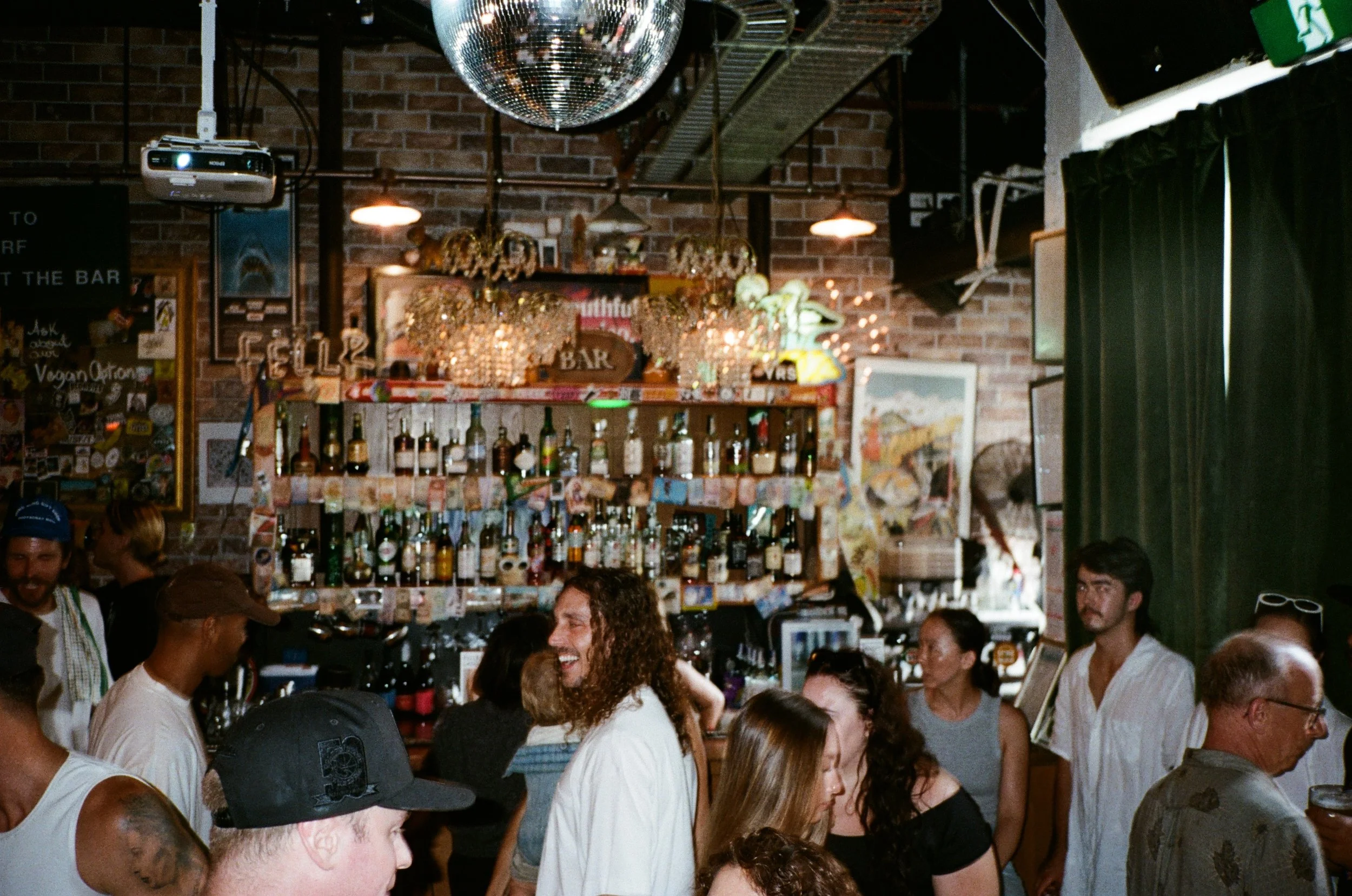 A lively bar scene with people socializing and enjoying drinks, with a brick wall background, bottles on shelves, a disco ball hanging from the ceiling, colorful flowers as decoration, and a chalkboard menu on the wall.