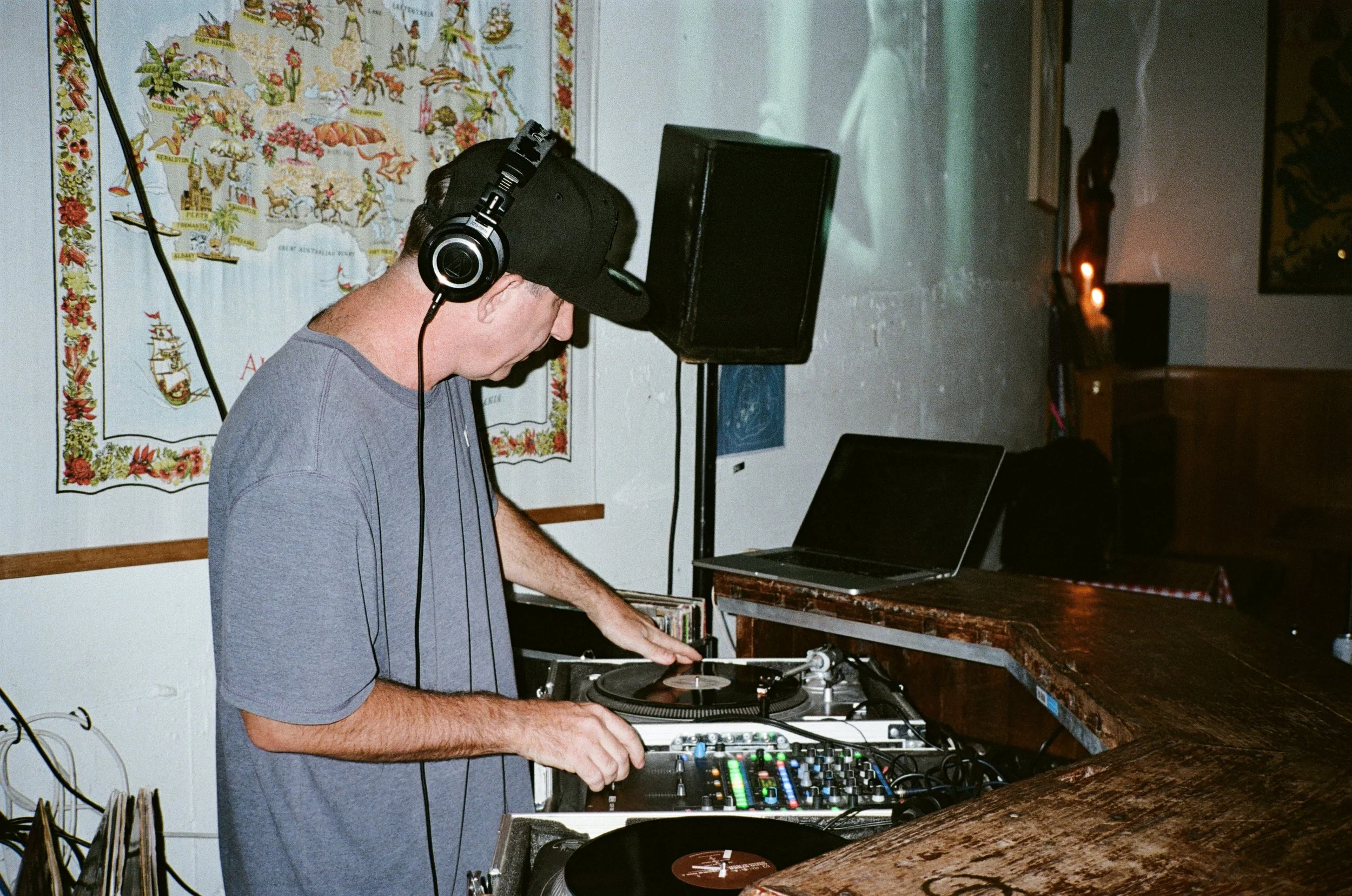 A man wearing headphones and a cap is DJing at a setup with turntables and a mixer in a dimly lit room with candles and artwork on the wall.