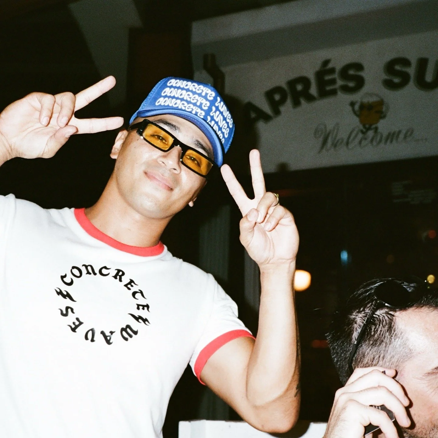Man wearing yellow-tinted sunglasses, a blue bandana, and a white T-shirt making peace signs with both hands at an indoor event.