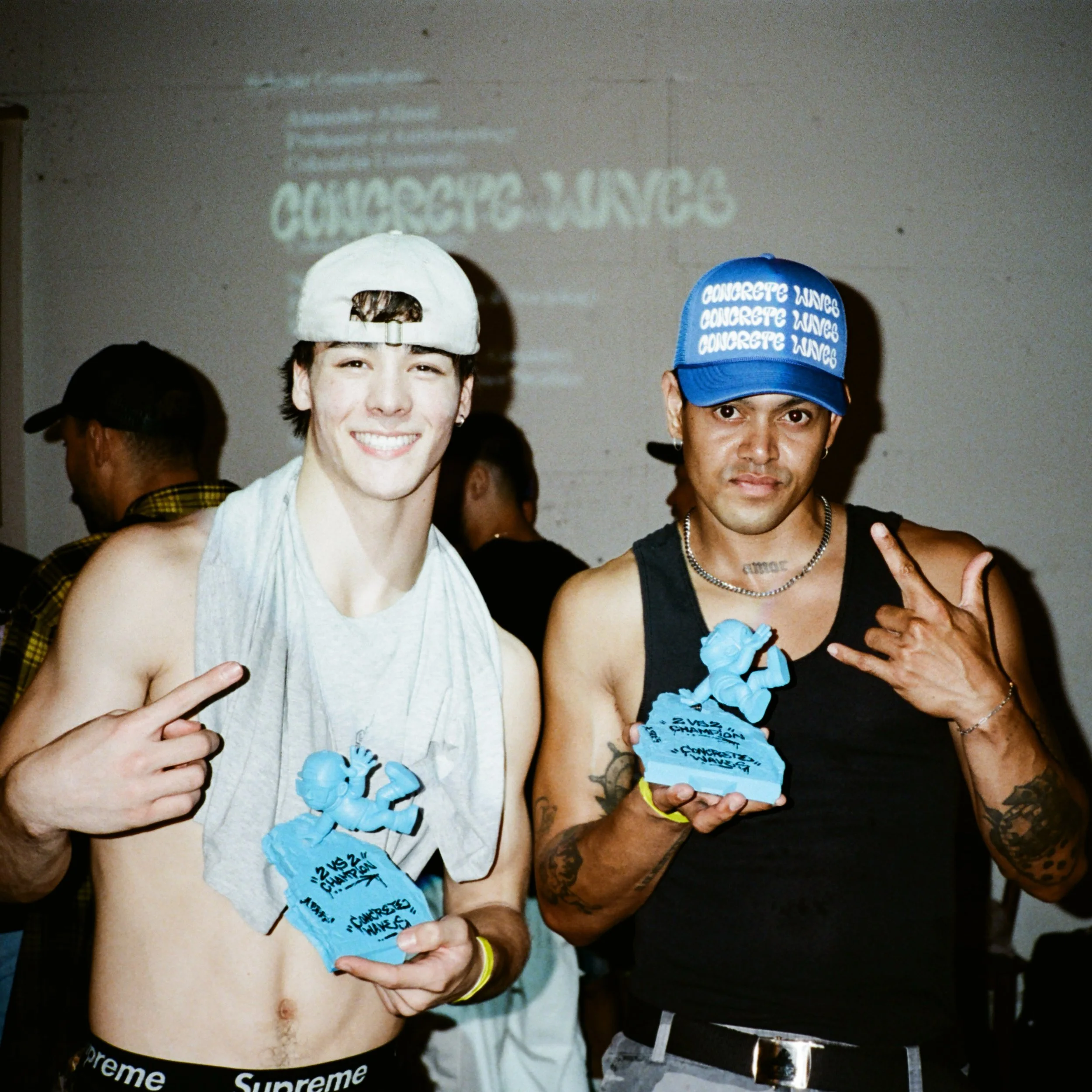 Two young men at a celebration with one showing a toothy smile, both wearing blue baseball caps with white text, holding blue cutouts, and making hand gestures. The background features a wall with a blurred reflection of a sign that reads 'CONCRETE WAVES'.