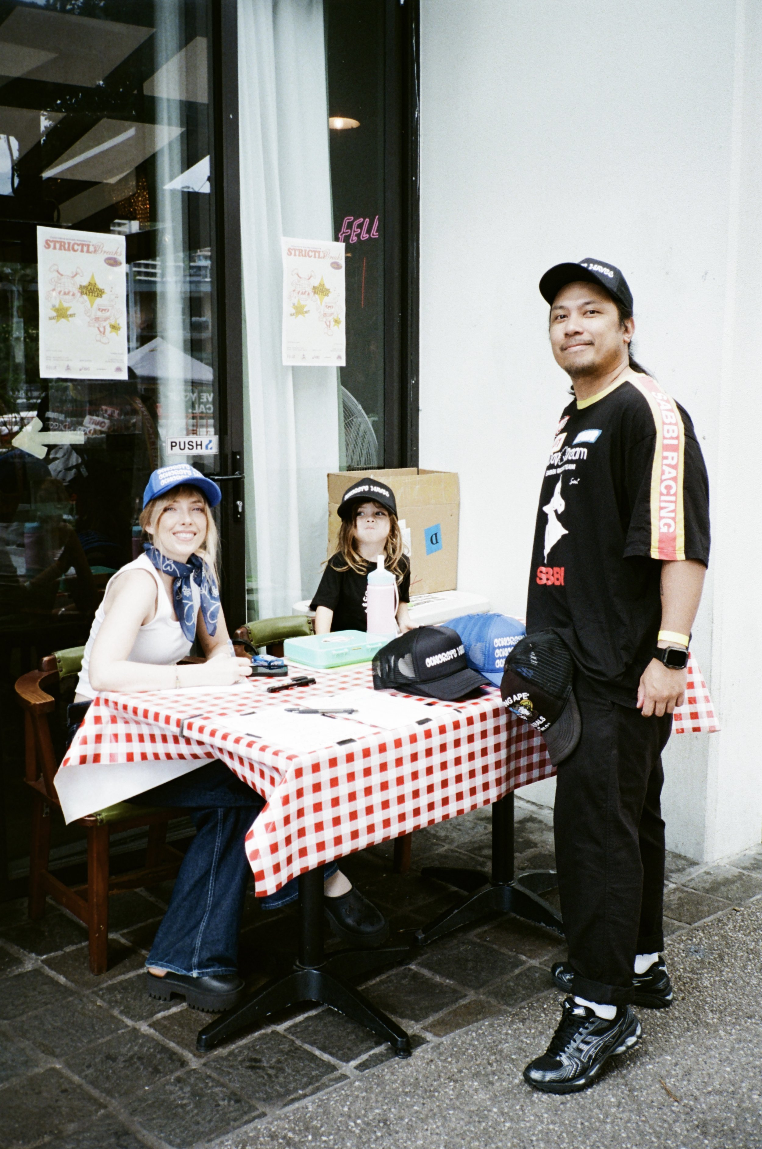 Three people gathered around a table with a red and white checkered tablecloth outside a restaurant. The woman seated on the left is smiling and wearing a blue cap, a white shirt, and a blue bandana. The woman in the middle has long hair, a black cap, and a black shirt. The man on the right, standing, is wearing a black racing-themed shirt, black pants, a black cap, and sports shoes.