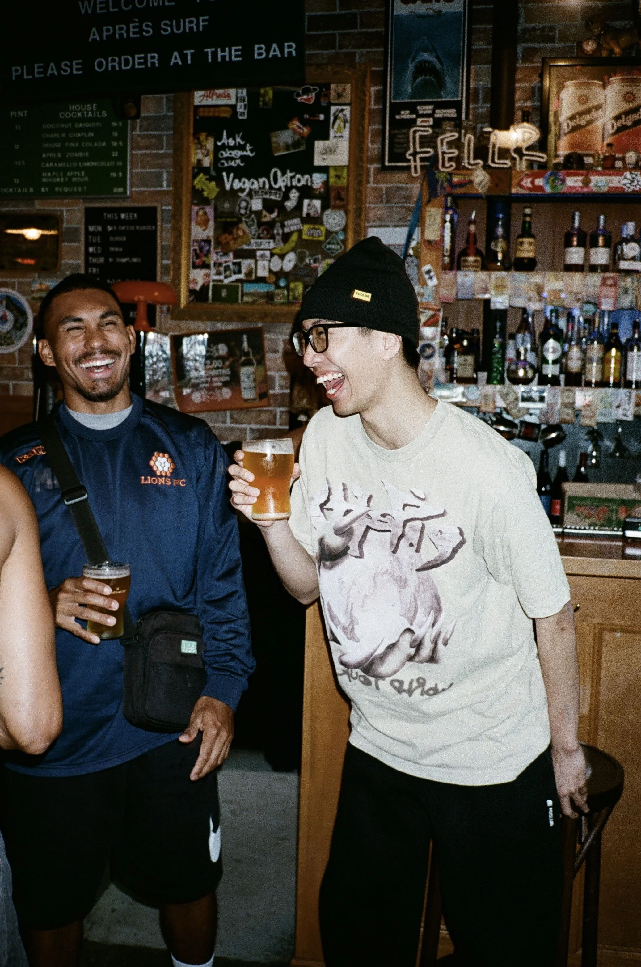 Two young men are smiling and holding drinks in a bar. One is wearing a blue sports jersey with 'LIONS FC' and a shoulder bag, and the other is wearing glasses, a black beanie with a yellow tag, and a graphic t-shirt.