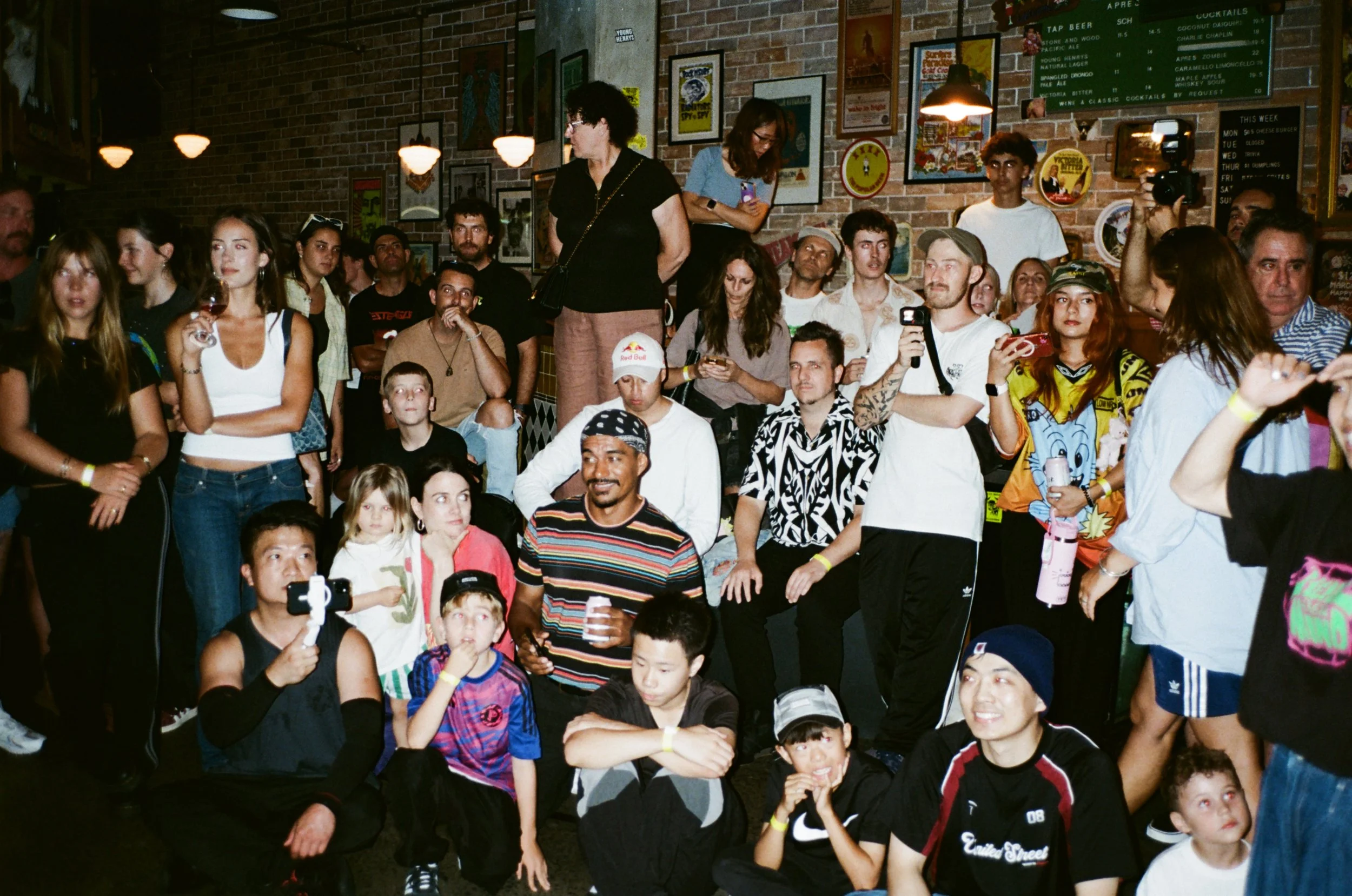 A group of people gathered indoors at a social event, with a brick wall decorated with posters and framed artwork in the background. Some are standing, others seated, appearing engaged or taking photos.