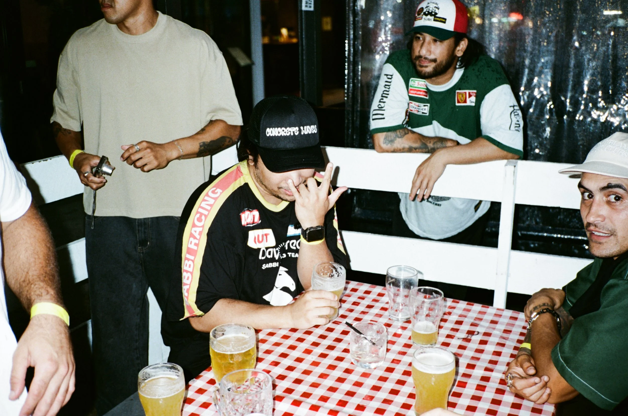 People at a gathering with glasses of beer on a red and white checkered tablecloth, some standing and some sitting, in a setting with a festive or casual atmosphere.