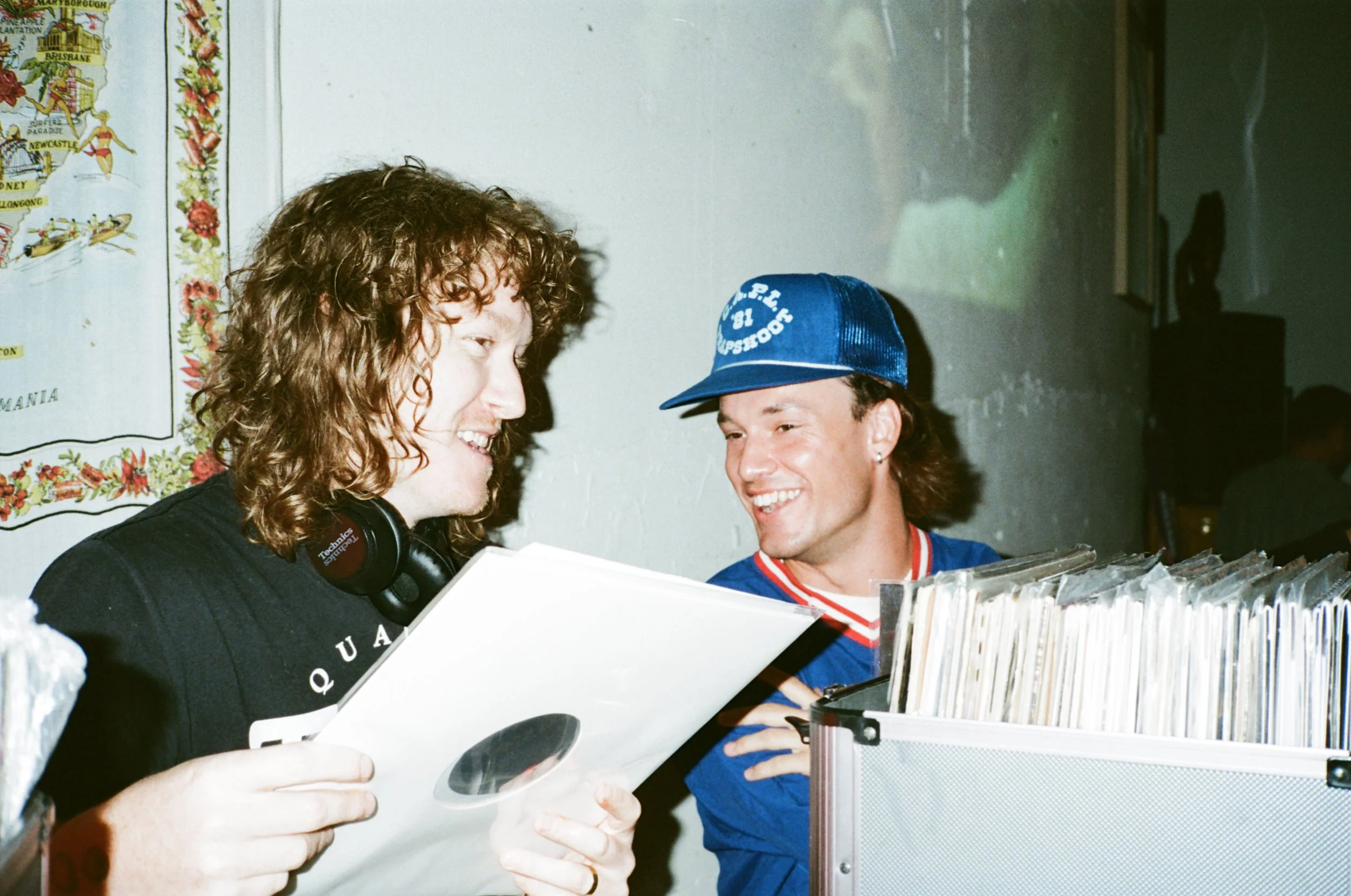 Two men with curly and straight hair look at records in a box, smiling, with one wearing an Ontario Blue Jays cap. They are indoors with a wall featuring a colorful map or poster in the background.