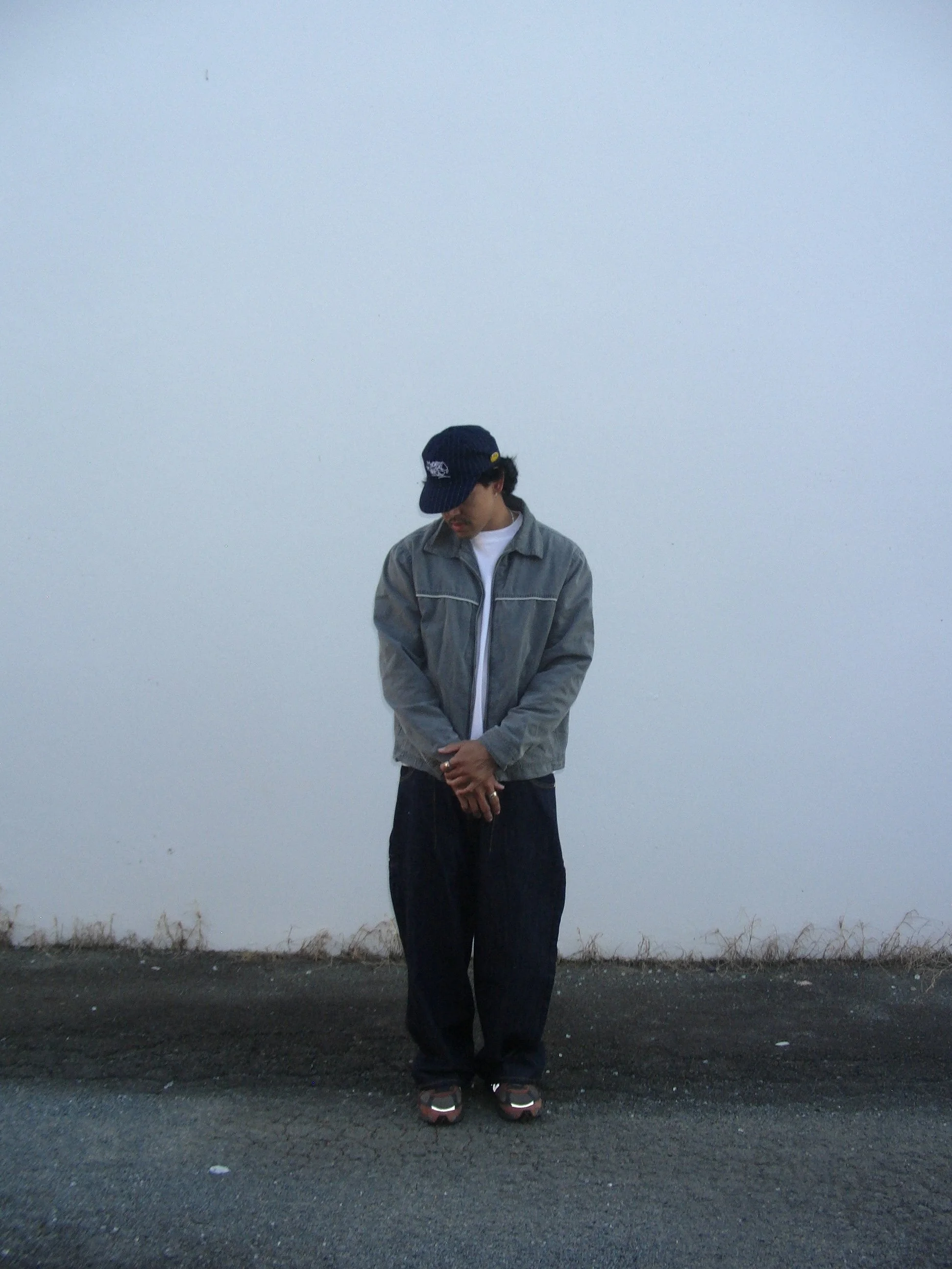 A person standing outdoors at dusk or dawn against a pale sky and a white wall, wearing a gray jacket, dark pants, and a black cap, with his head slightly bowed and hands clasped.
