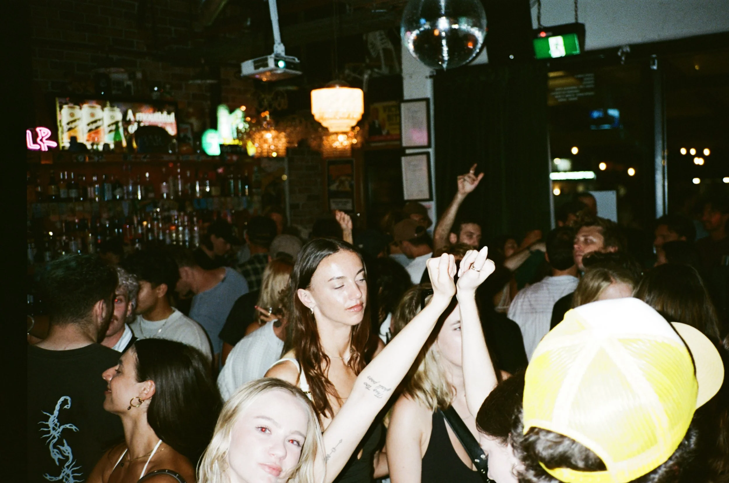 A crowded nightclub with people dancing and socializing. The background shows a bar with bottles, a television screen, and colorful lights. The scene is lively and energetic, with a disco ball hanging from the ceiling.