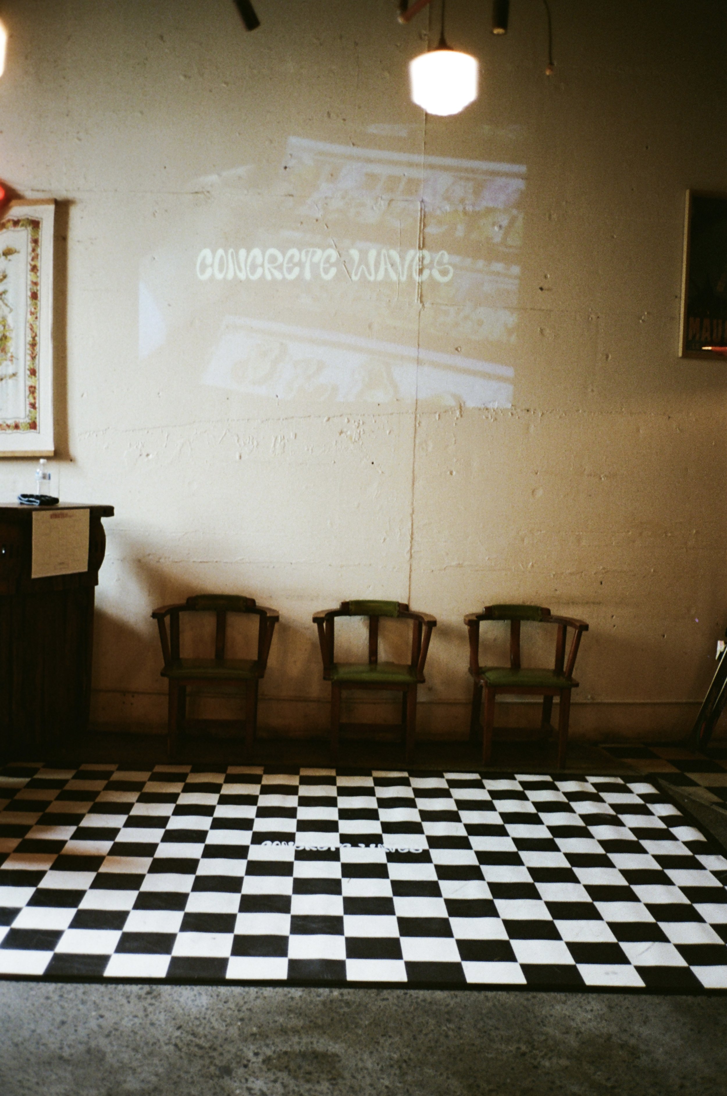 A wall with the words 'CONCRETE WAVES' projected on it. Below the wall, there are four wooden chairs with green cushions arranged in a row. The floor has a black and white checkered pattern, and there is a small table to the left with a bottle of water on top.