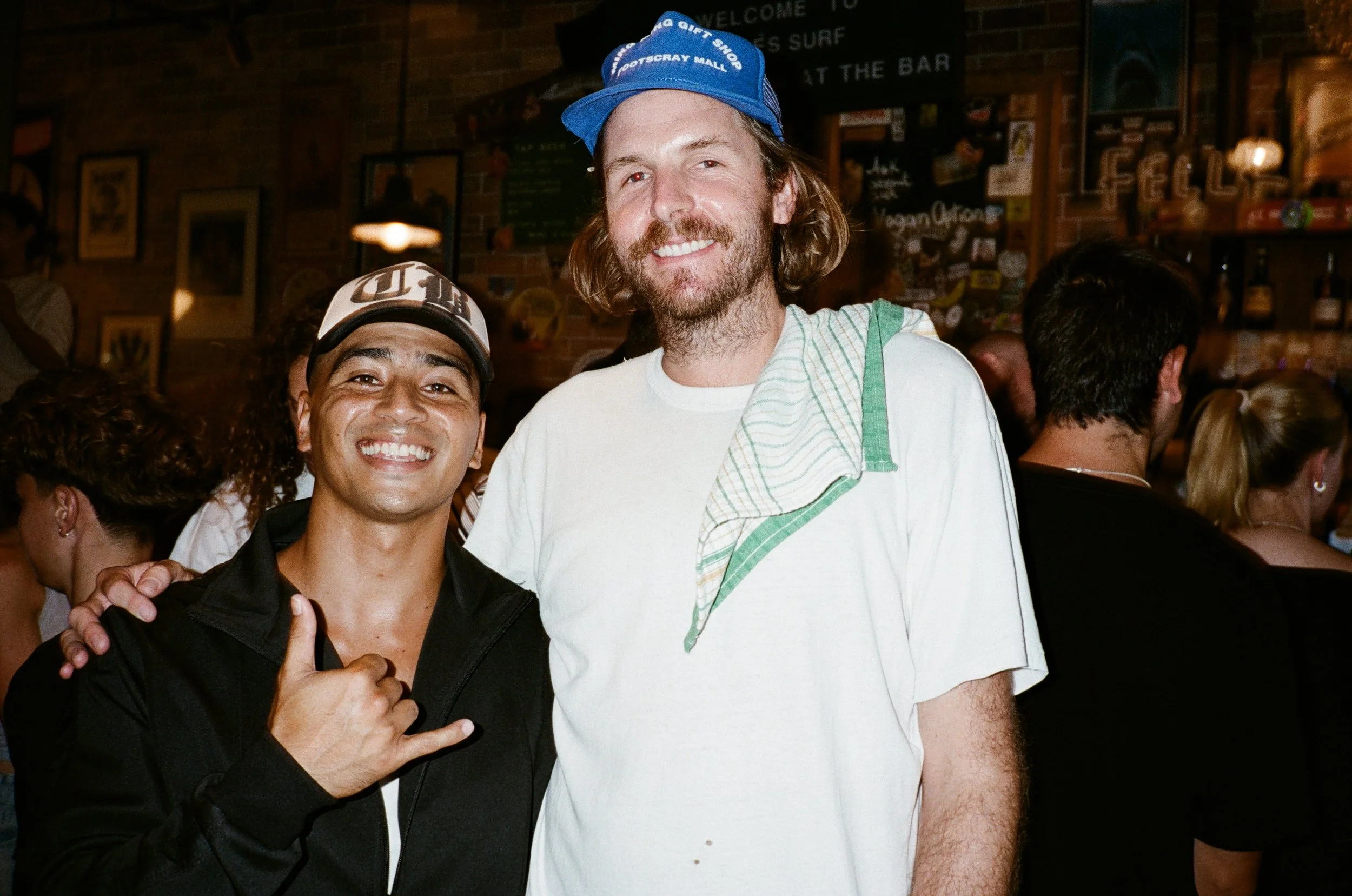 Two smiling men in a bar or pub, one wearing a black jacket and a cap, the other in a white t-shirt with a green towel over his shoulder. The man in the cap is making a hand gesture resembling a shaka sign.
