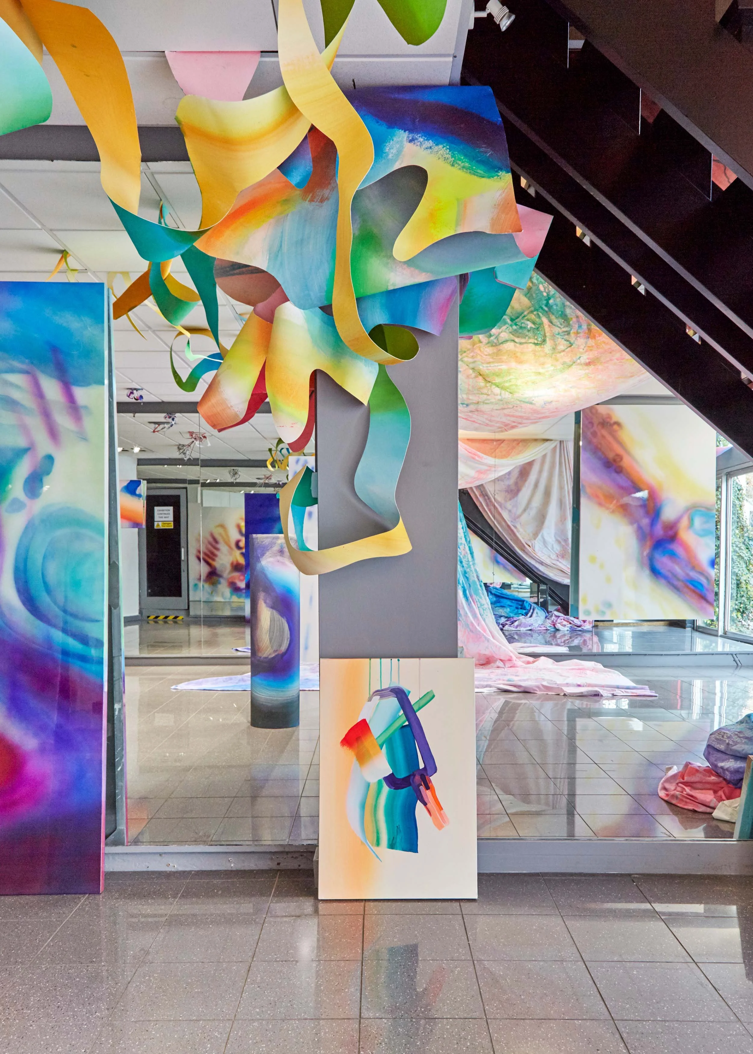 Colorful abstract art installation with hanging paper-like sculptures and vibrant paintings in a gallery setting.