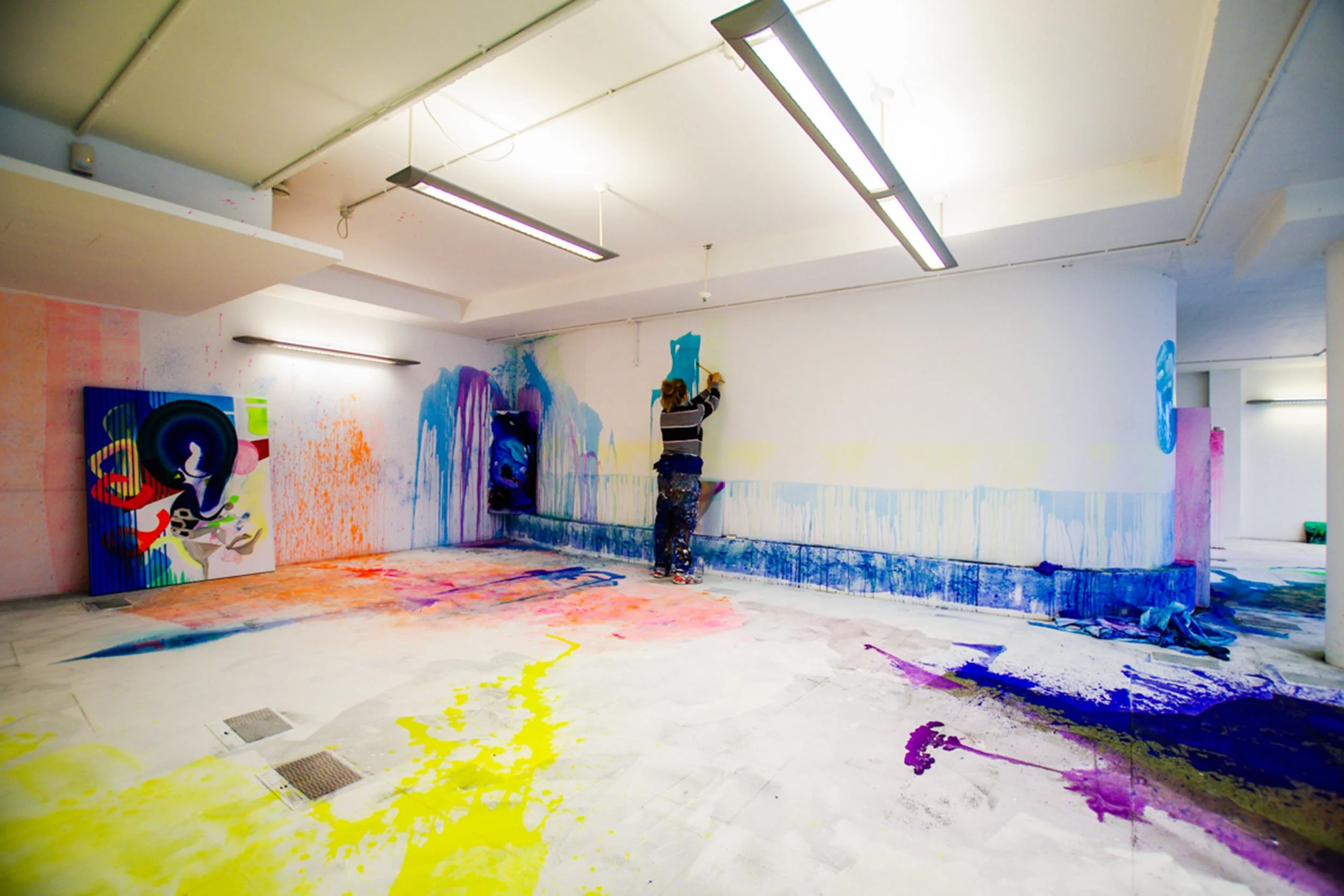 Person creating colorful abstract graffiti art on a large white wall in an indoor space, with various splashes of paint on the floor.