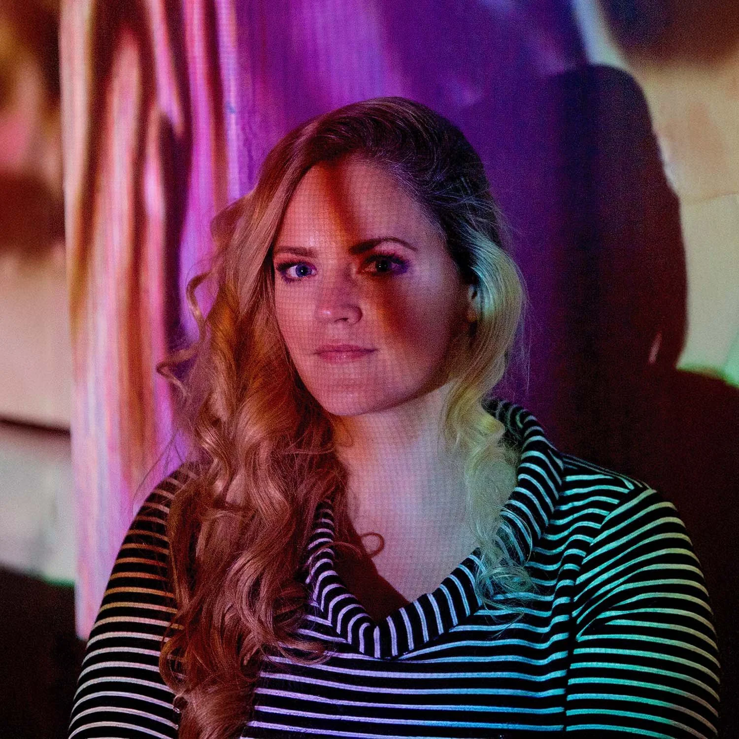 A woman with long, wavy blonde hair, looking at the camera with a neutral expression, wearing a black and white striped top, standing against a colorful background with pink and purple lighting.