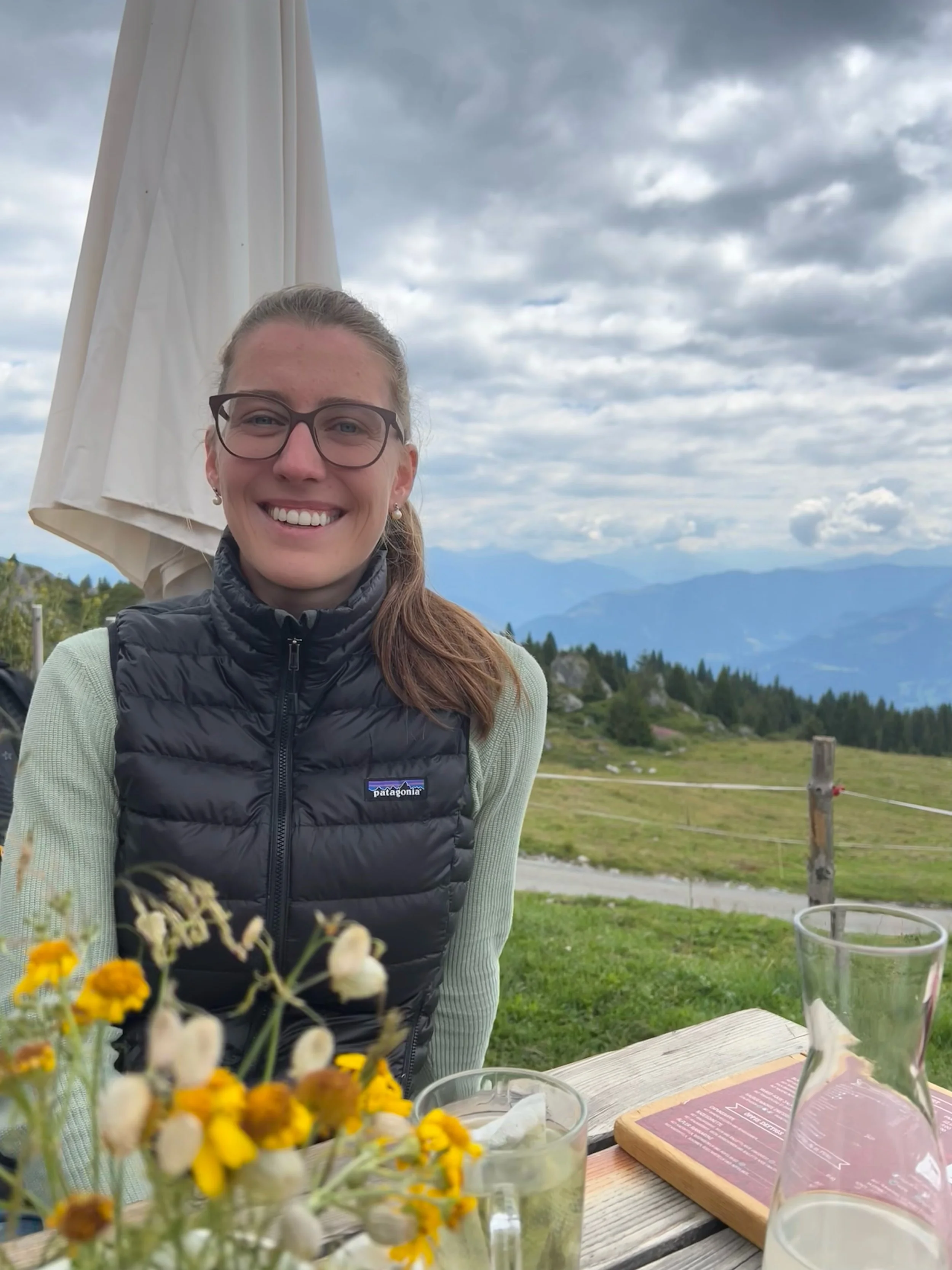 Alina Lea Moser mit Brille und grauem Pullover sitzt vor einem Bergpanorama, lächelt. Es regnet leicht, Wolken teilen die Berge, im Vordergrund Blumen und ein Tisch mit Getränken.
