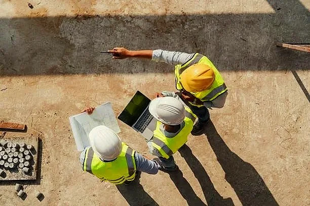 Three construction workers wearing safety vests and helmets looking at a laptop on a construction site.