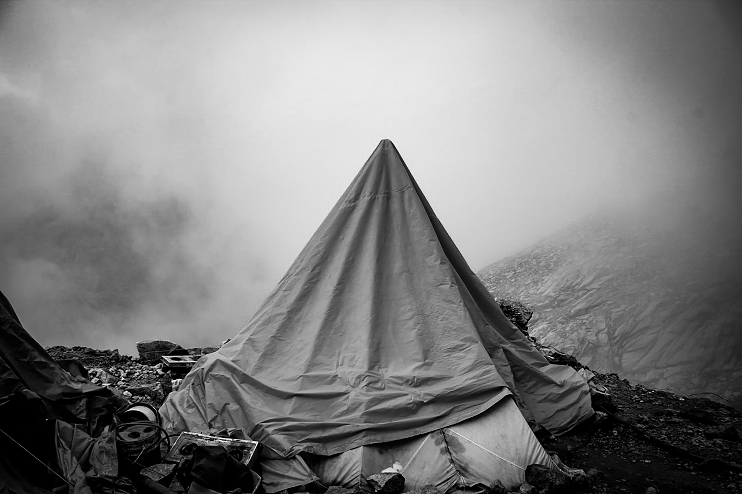 A wilderness campsite with a conical tent on rocky terrain, surrounded by fog or clouds.
