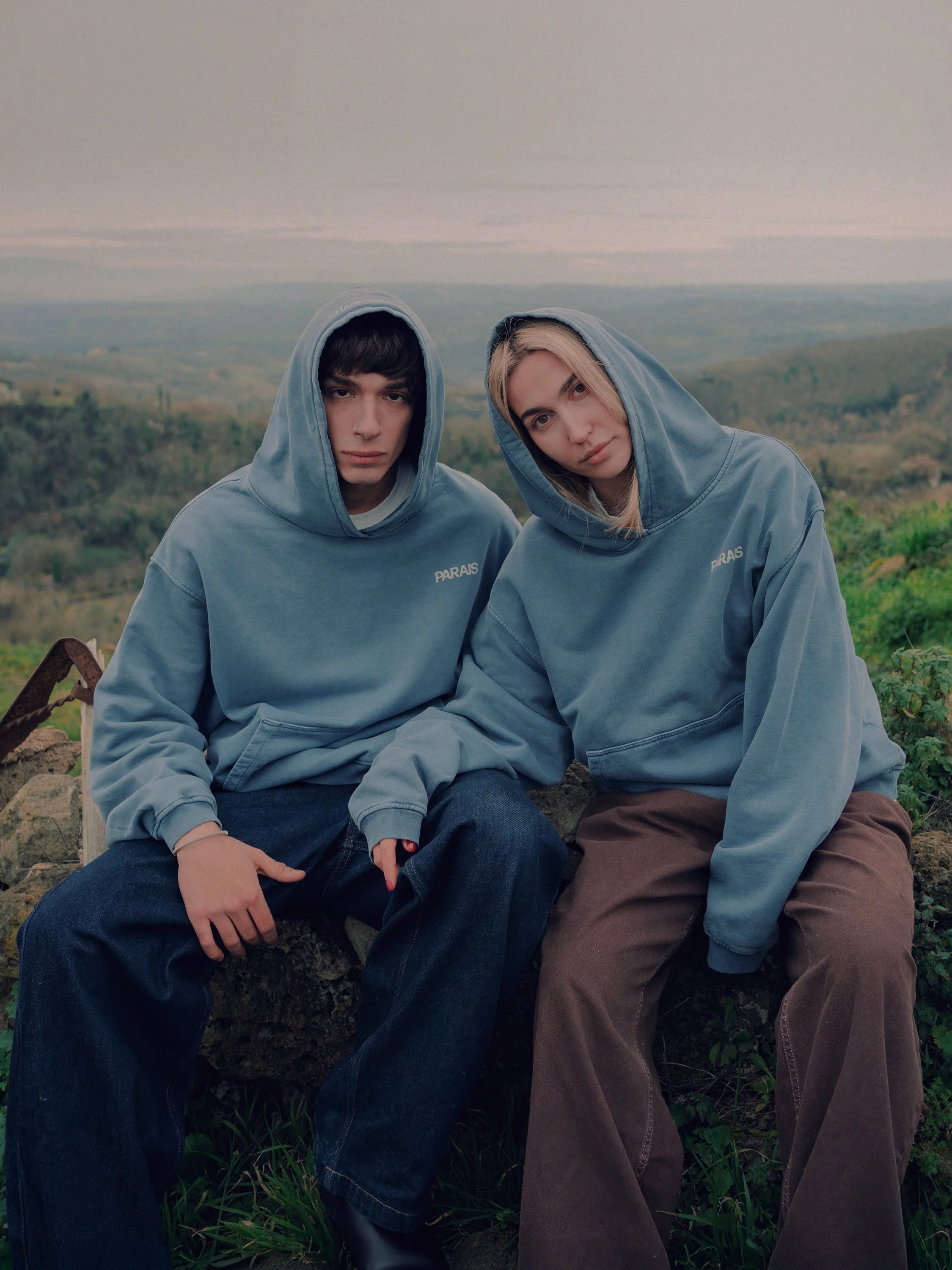 Two young people sitting outdoors on a rock, wearing matching blue hoodies with the word 'PARAIS' on them, with a scenic landscape in the background.