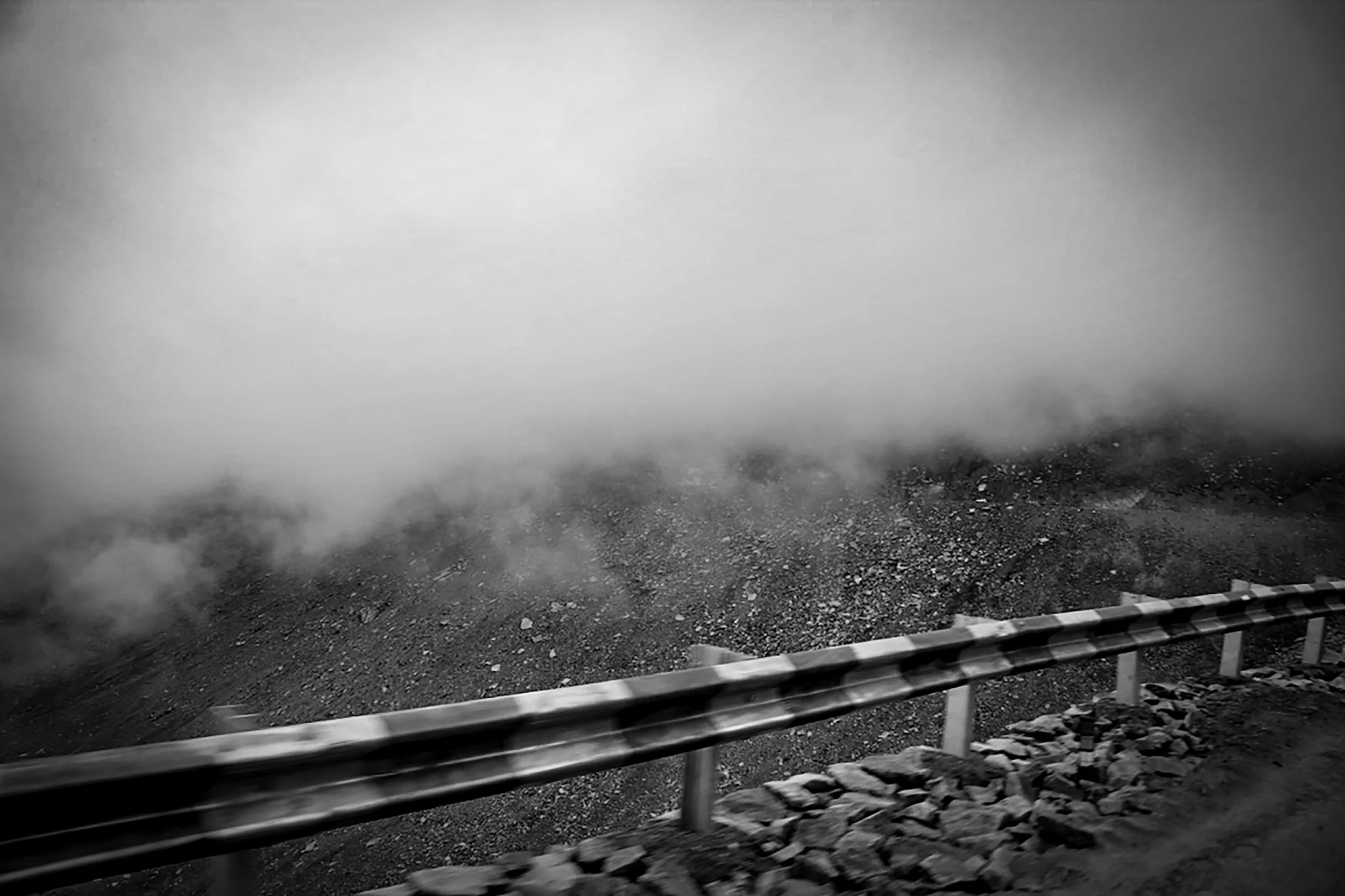 Black and white photo of a mountain landscape with a guardrail along a road. Fog or clouds obscure the mountain peak in the background.