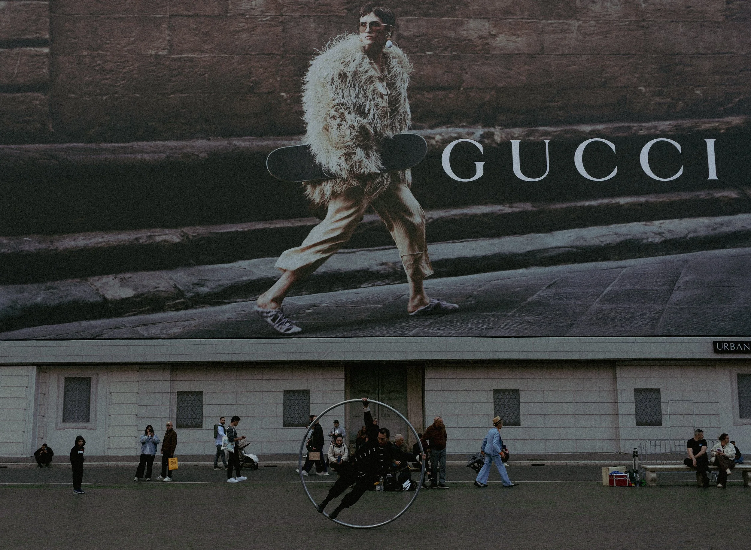 A large billboard showing a person walking with a skateboard in front of the Gucci logo. Below the billboard, there are people walking, sitting, and standing in an outdoor area, with some holding bags and cameras, and a performer in a circular hoop p
