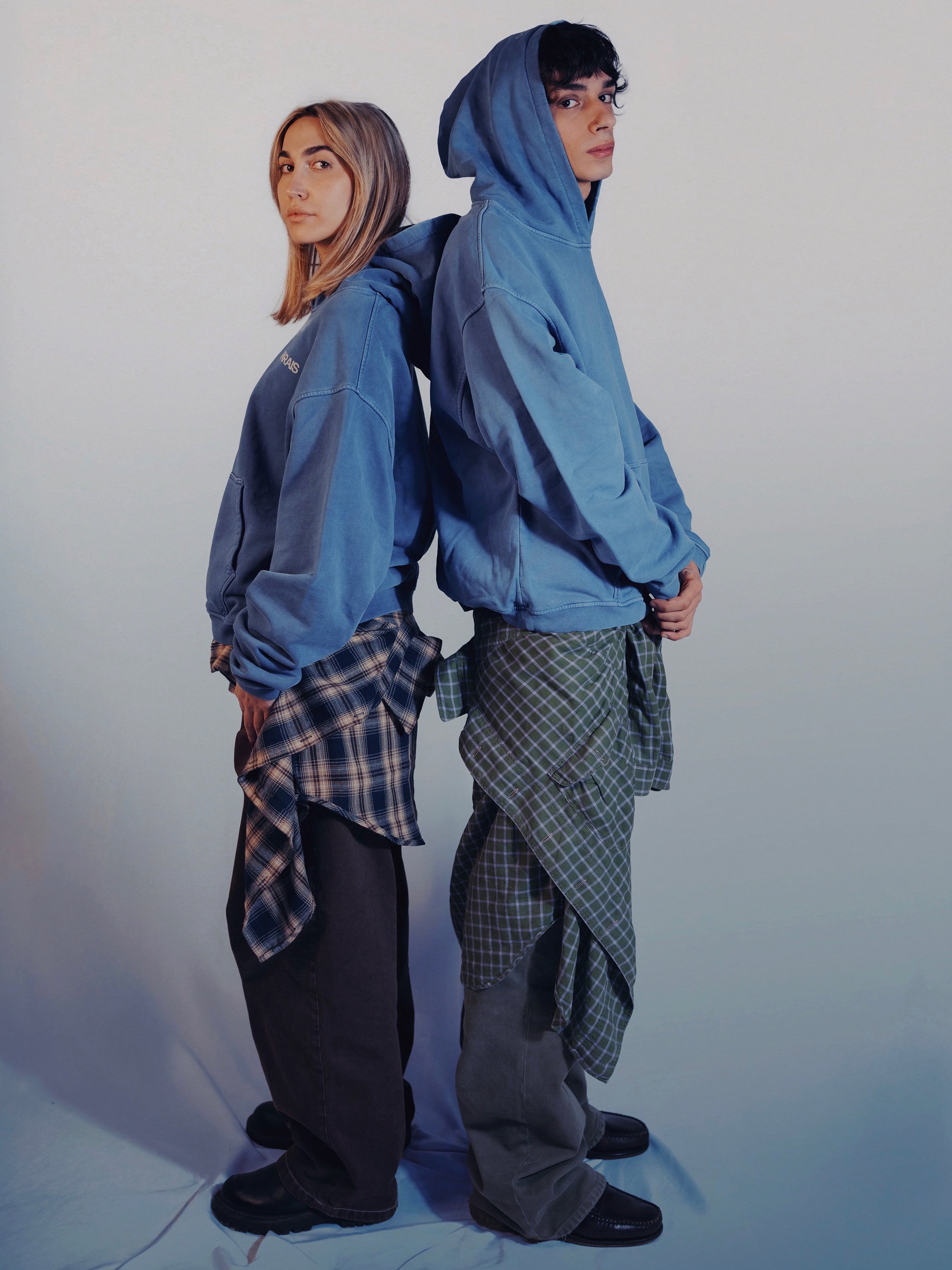 Two young people, a woman and a man, stand back-to-back against a plain light-colored background, both wearing blue hooded jackets and plaid shirts tied around their waists.