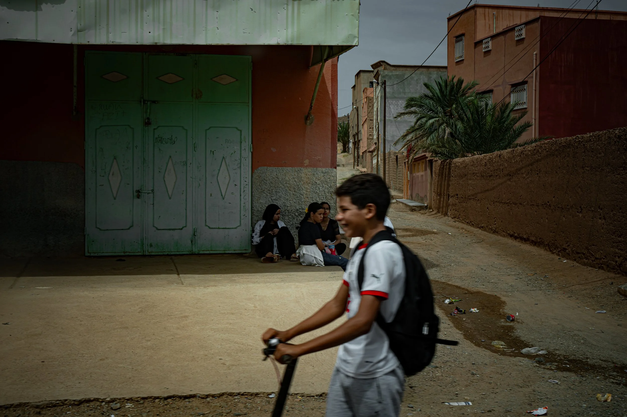 A young boy riding a scooter on a dirt road in a neighborhood with multiple women sitting on the sidewalk against a green door, and houses and palm trees in the background.