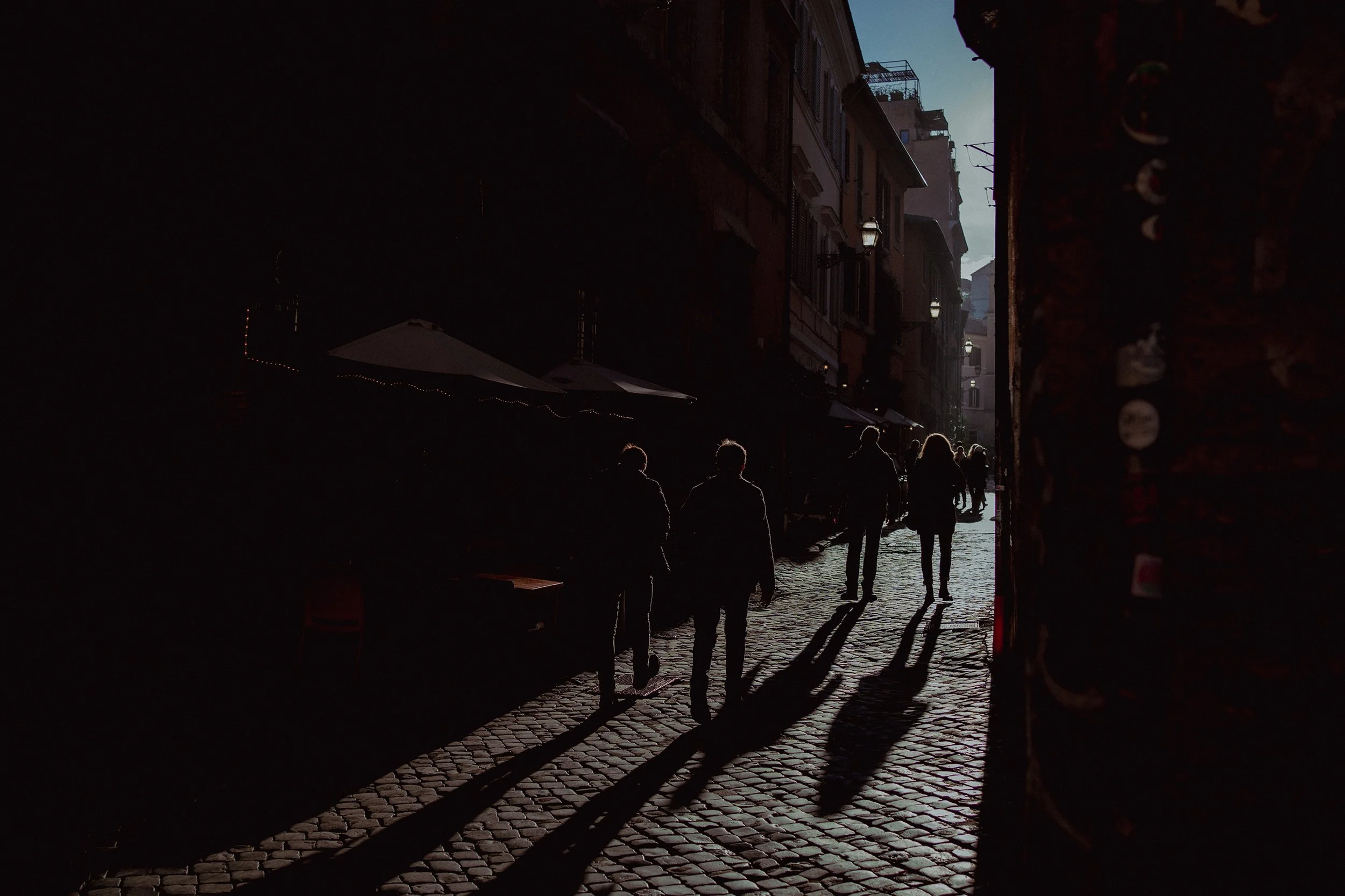 People walking on a cobblestone street in silhouette with sunlight creating long shadows, lined with buildings and outdoor umbrellas.