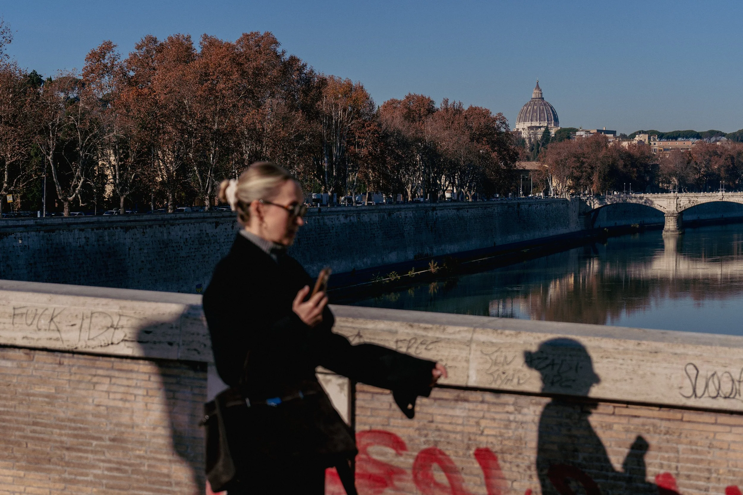 A woman with blonde hair tied back, wearing glasses, a black jacket, and holding a phone, standing on a bridge over a river. In the background, trees line the riverbank, and a large domed building is visible under a clear sky.