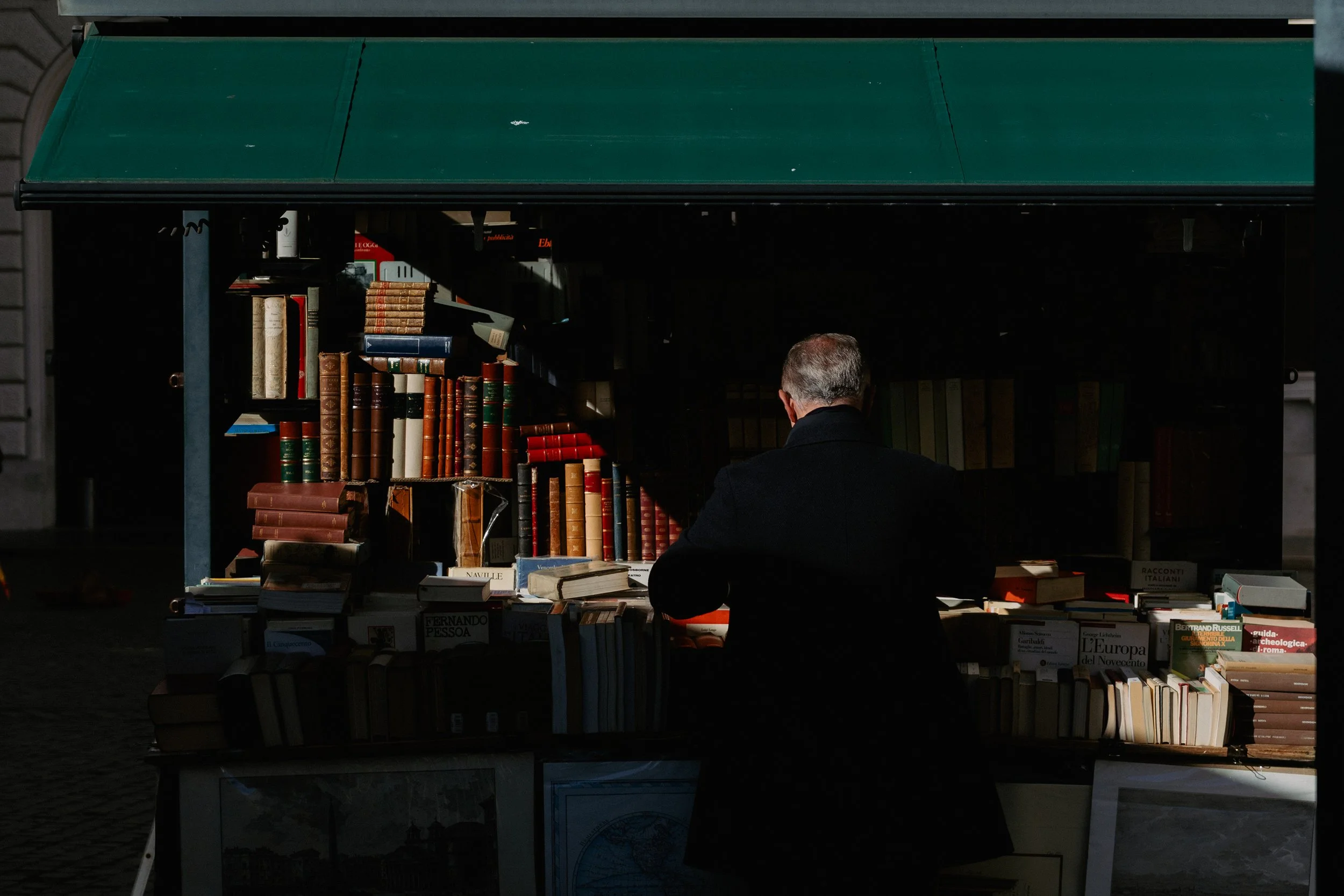 A person with gray hair wearing a black coat browsing books at an outdoor book stall under a green canopy.
