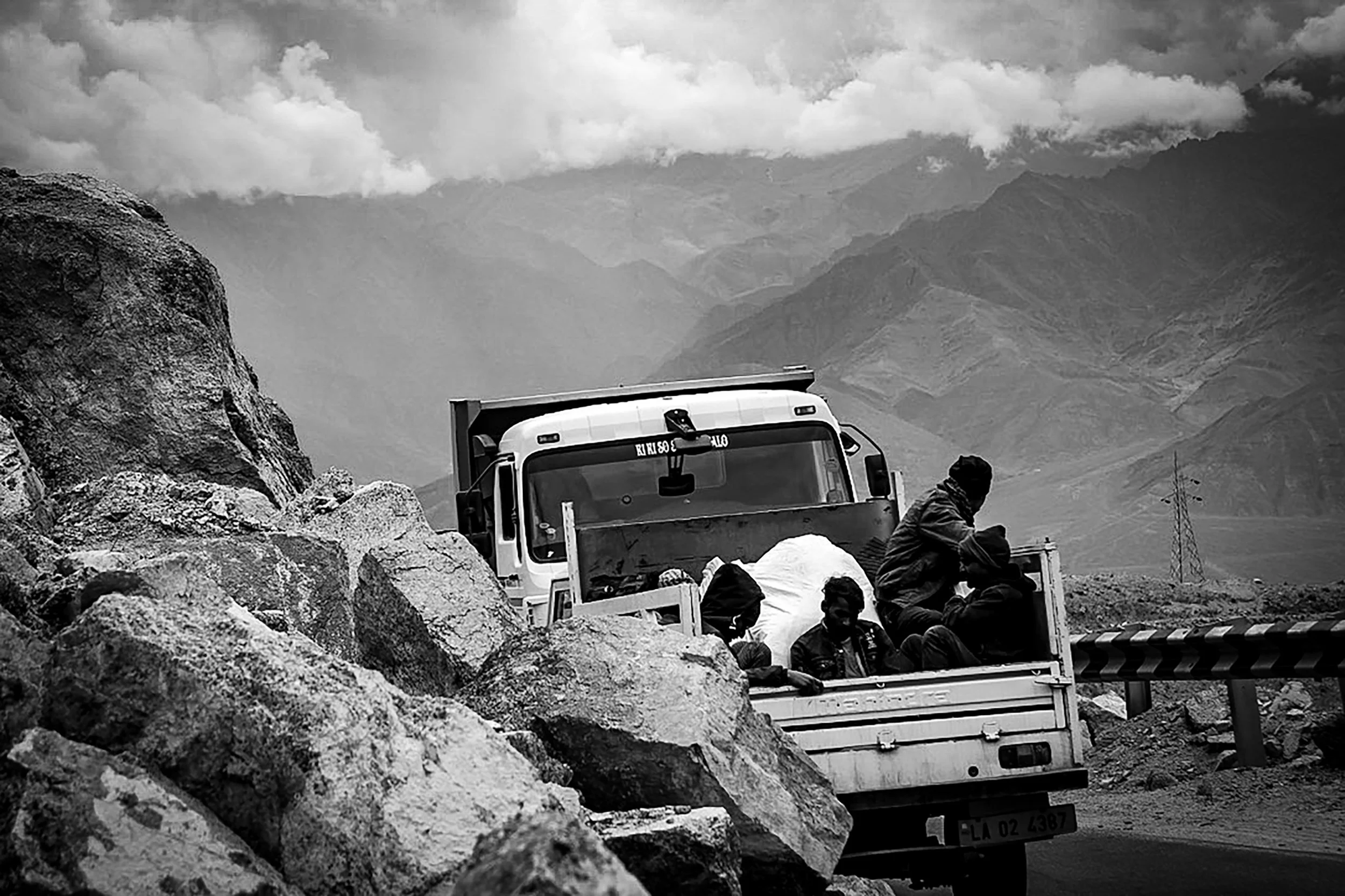 A truck is traveling on a mountain road with large rocks on the side. There are three people in the truck's cargo bed, sitting and relaxing. Mountainous terrain and cloudy sky are in the background.