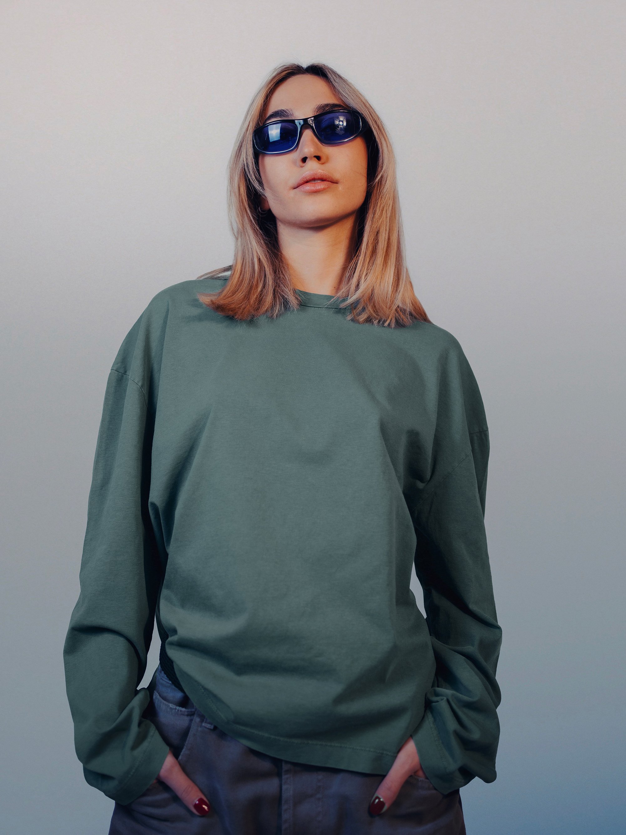 A woman with blonde hair wearing dark sunglasses, a loose olive green shirt, and gray pants against a plain light background.