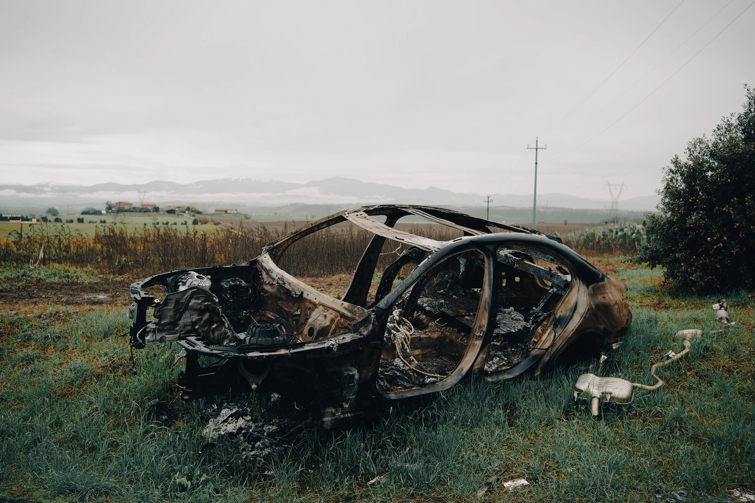 Burned-out car wreck in a grassy field with a cloudy sky and distant mountains.