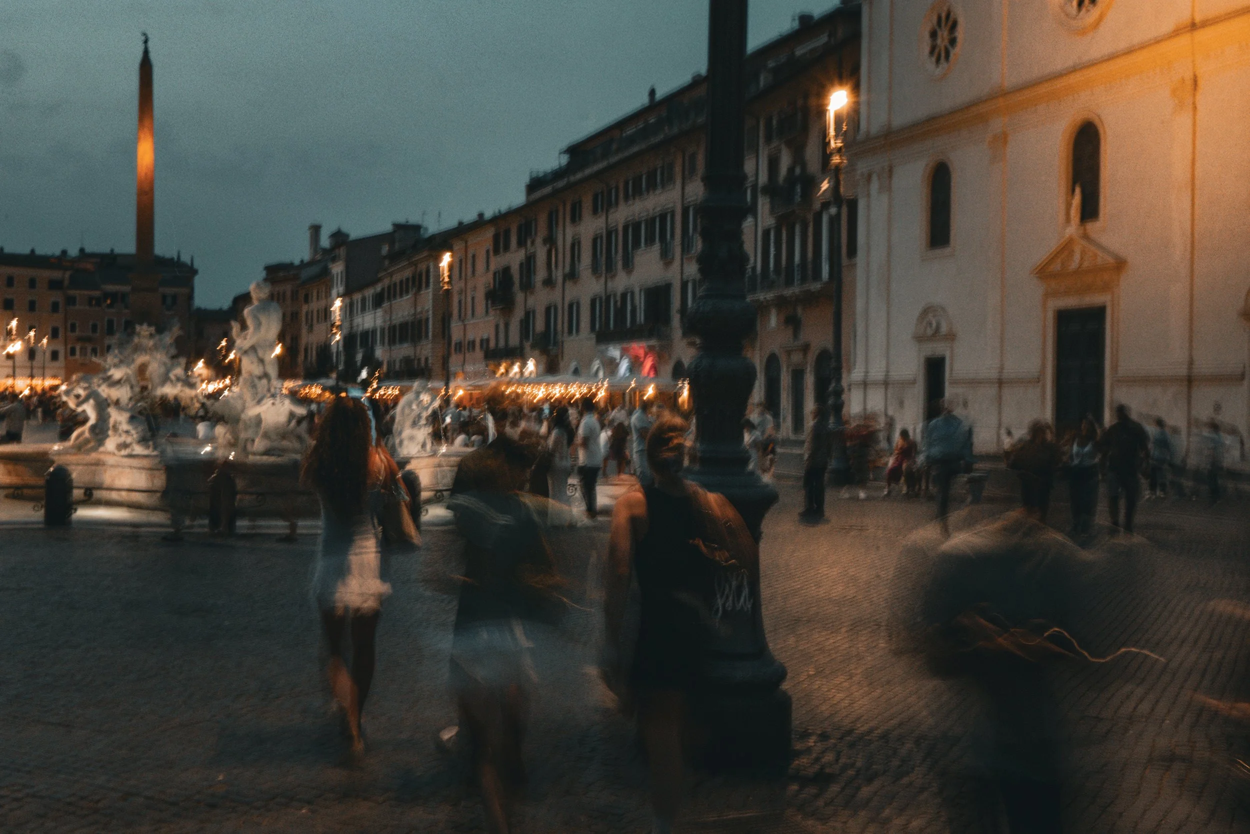 A lively city square at dusk with a fountain, historic buildings, and many blurred pedestrians walking around.