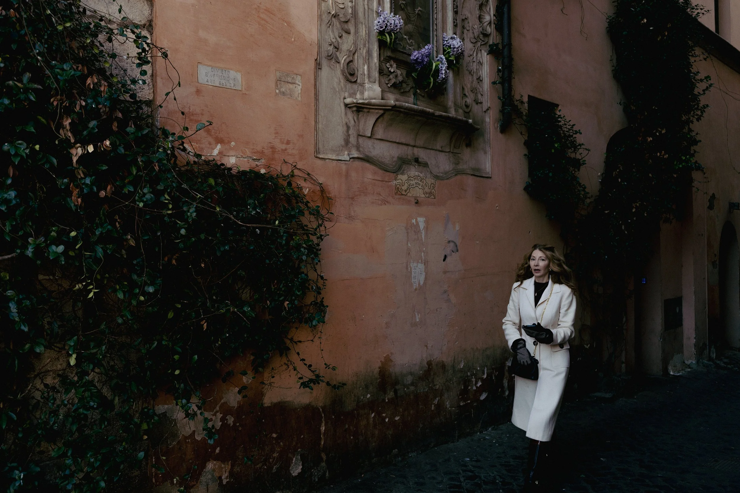 A woman in a white coat and black gloves walking along a narrow cobblestone street with old pink buildings and greenery, possibly in a European city.